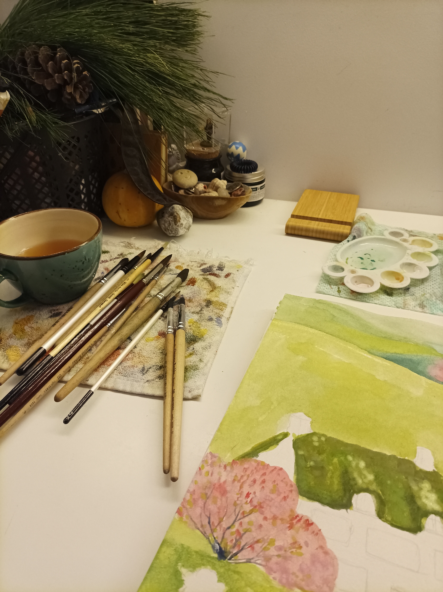 The image displays an indoor art workspace located in Batumi, Georgia. The scene is presented from a slightly elevated perspective, looking down onto a light-colored desk surface.
In the foreground, to the left, a teal-colored ceramic mug with an unidentifiable amber liquid rests on the desk. Adjacent to it, a collection of approximately two dozen paintbrushes, varying in size and type, with wooden handles and synthetic bristles, are laid across a small, light-colored fabric towel stained with multiple colors. To the right of these, a white plastic watercolor palette, containing visible pigments including green, yellow, and white, is situated on another stained fabric cloth. A sheet of watercolor paper occupies the lower right portion of the frame, showcasing a partially completed landscape painting. This painting features a pink-flowered tree on the left, foregrounded by green, and a background depicting green hills or foliage with unpainted white areas suggesting unfinished architectural or structural elements.
In the midground, behind the mug and brushes, a dark, open-weave container holds several pine branches with needles and two large pinecones. To its right, a small, round orange gourd and a dark, elongated seed pod are present. A small wooden bowl contains multiple light-colored, irregularly shaped items, one of which exhibits a faint smiley face. A small, textured grey stone lies beside the bowl. Further right, behind the wooden bowl, various small containers and objects are partially visible, including a dark jar with a black lid and a blue and white patterned spherical object. A small, light-colored wooden block or stand is positioned to the left of the paint palette.
The background consists of a plain, light-colored wall. No individuals are present in the scene. The arrangement of materials indicates an active or recently utilized area for watercolor painting.
