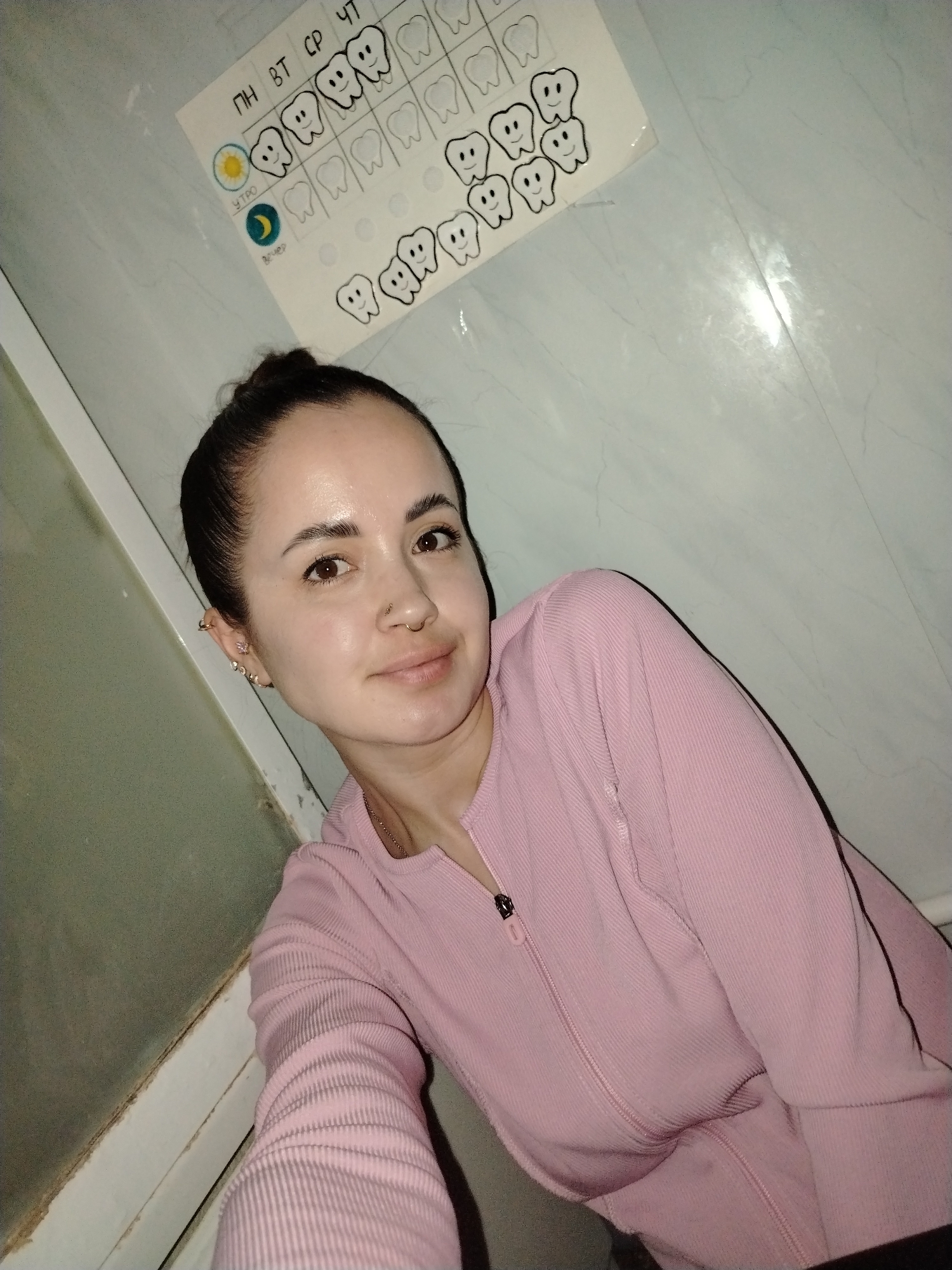 A selfie-style photograph shows a woman in a pink zip-up shirt. Her hair is pulled back in a bun, and she has multiple ear piercings and a nose ring. A white tiled wall is visible behind her, and a chart with teeth and Russian letters is adhered to the wall. The location is Noviy, Russia.