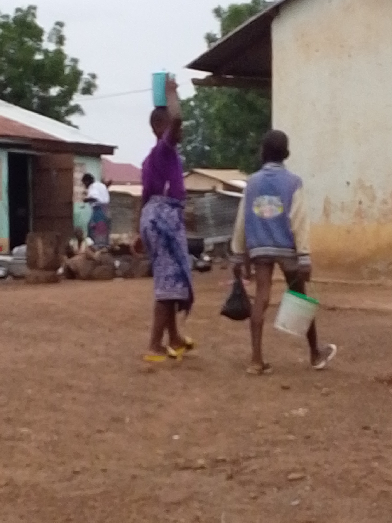 The image captures an outdoor scene, possibly in a village or rural setting, during daylight hours. The ground appears to be dirt or packed soil.
Two children are the main subjects. One child, wearing a purple shirt and a patterned blue and white cloth wrapped around the waist, is walking with a blue container balanced on their head. The second child is wearing a blue and cream-colored jacket and shorts, carrying a black plastic bag in one hand and a white bucket with a green rim in the other.
In the background, there is a simple building with a corrugated metal roof. A woman, dressed in a white top and a patterned skirt, is visible in the doorway. There are also piles of sacks or bundles near the building. Another building is visible behind the first. A tree is situated behind the building, adding to the natural backdrop.