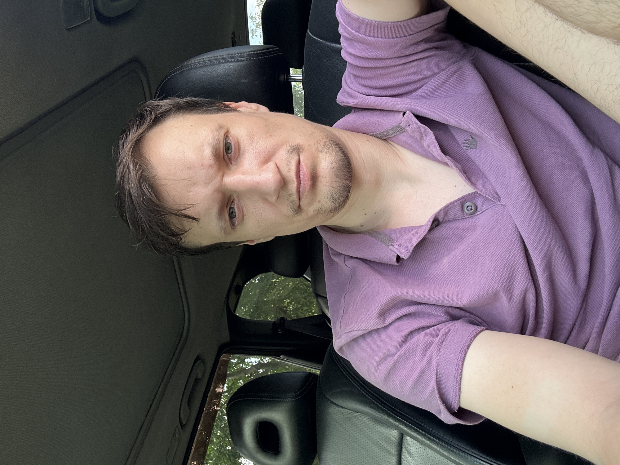 A man is sitting in the driver's seat of a car, likely in Makiv, Ukraine. He has short, dark hair and is wearing a light purple polo shirt. The interior of the car is visible, including black leather seats and a sunroof. Trees and foliage can be seen through the car windows, suggesting an outdoor setting. The man is looking directly at the camera with a neutral expression.