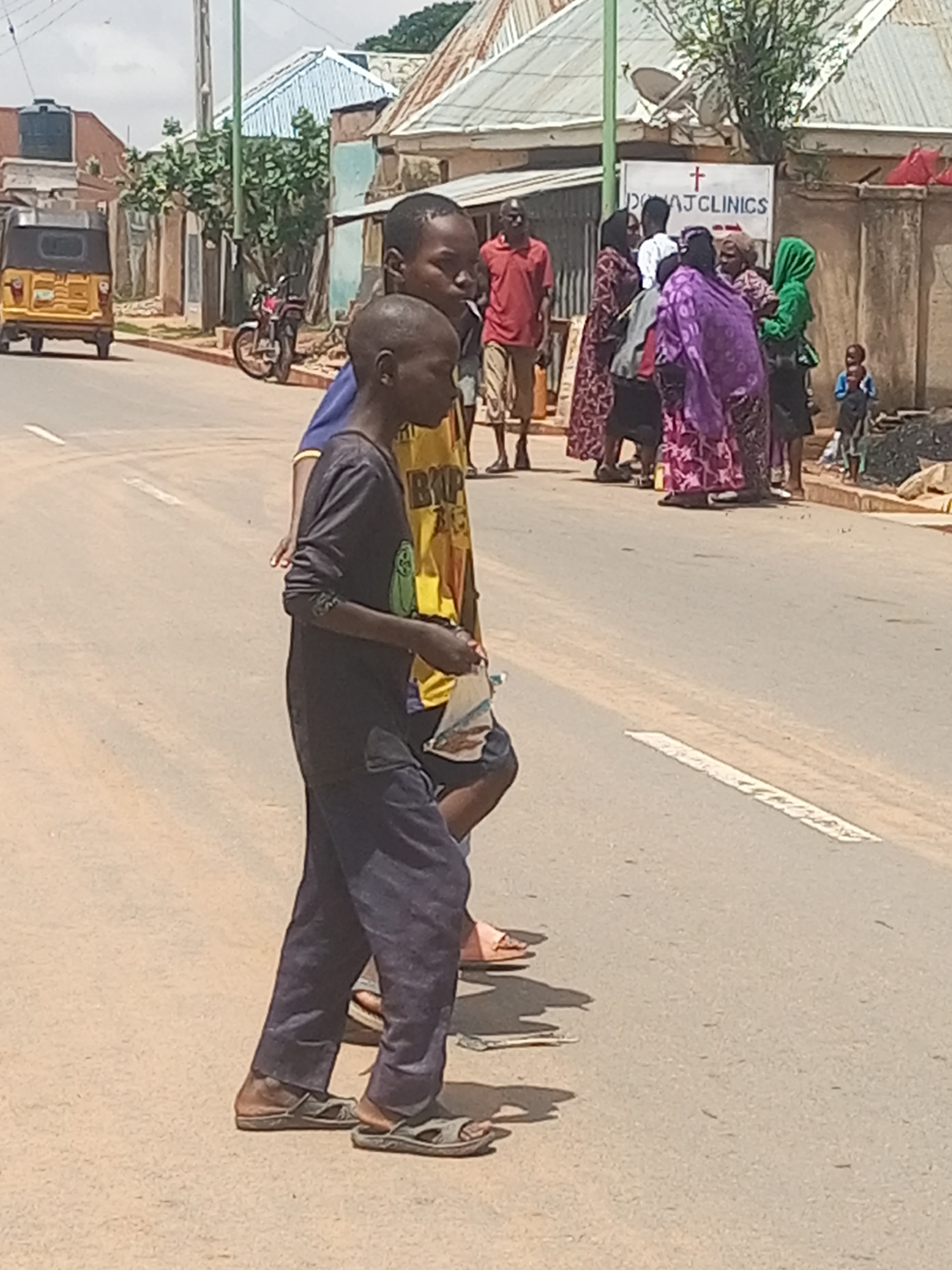 In Gombe, Nigeria, a street scene unfolds under a bright sky. Two young boys, one in a dark long-sleeved shirt and dark pants, the other in a yellow shirt with "BIG" printed on it, walk across an asphalt road. The boy in the dark shirt wears dark sandals, while the boy in the yellow shirt is only partially visible, wearing sandals. They are near a white line painted on the road.
To their left, a yellow and black motorized tricycle is parked on the side of the road. Further back, a red motorcycle is visible.
On the right side of the street, a group of people are gathered. A sign with a red cross and "DONAJ CLINICS" is affixed to a building. Several women, some wearing brightly colored headscarves and wrappers, are interacting. One woman in a green headscarf is particularly noticeable. A man in a red polo shirt and shorts stands to the left of this group. A small child in a blue shirt is also present, standing near some debris.
The environment suggests a bustling urban or semi-urban area with buildings lining the street and vegetation visible in the background. The overall impression is one of everyday life occurring on a street in Gombe.
