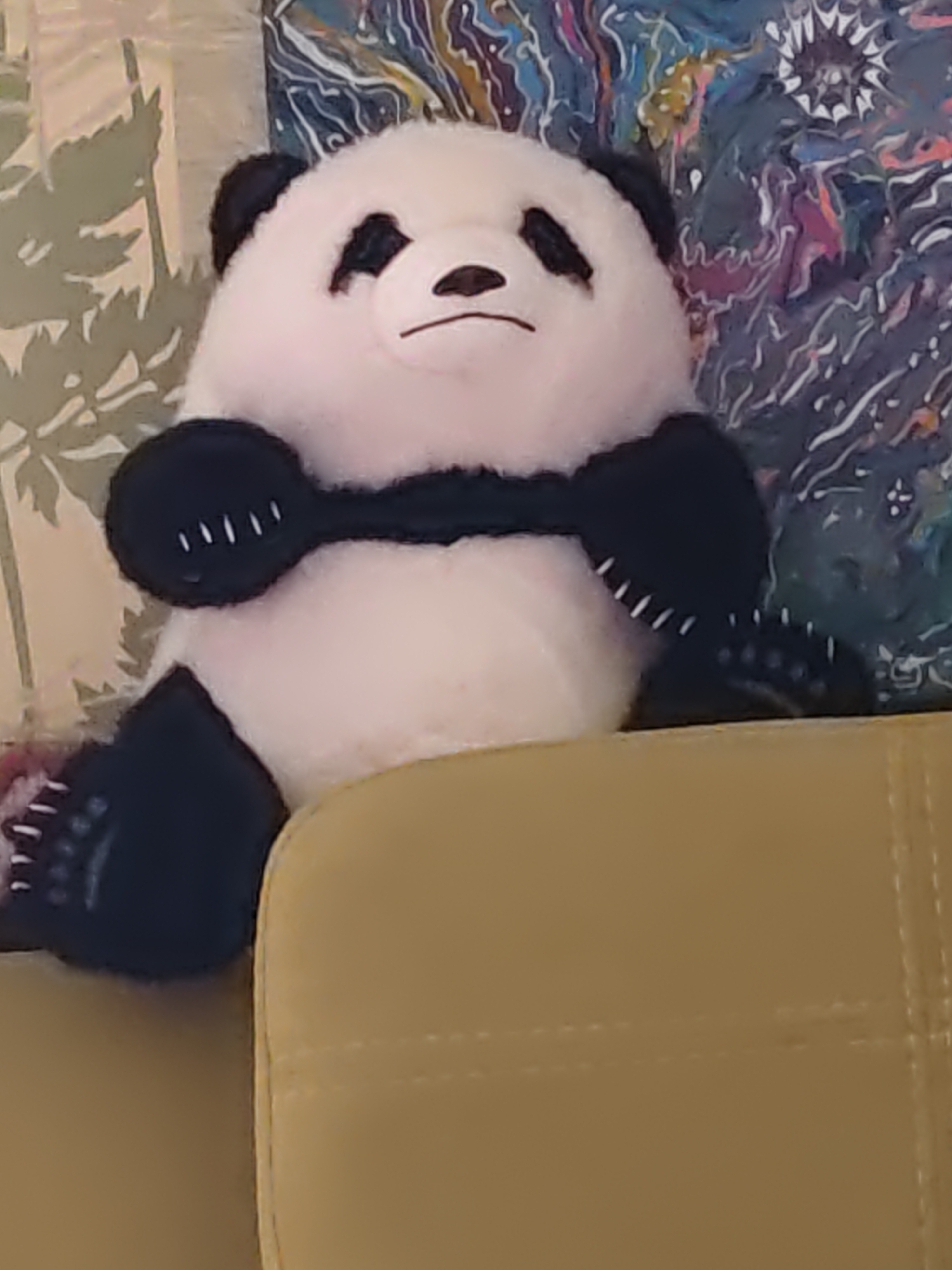 A close-up shot shows a stuffed panda bear sitting on a beige sofa. The panda is primarily white with black markings around its eyes, ears, and paws. It has a black band across its chest, resembling a scarf or a vest, with white stitching. The background features a tapestry or painting with abstract, colorful patterns in blues, purples, and yellows, and a faded green pattern with leaf-like shapes. The scene is indoors, with no indication of the city of Olginskaya, Russia.