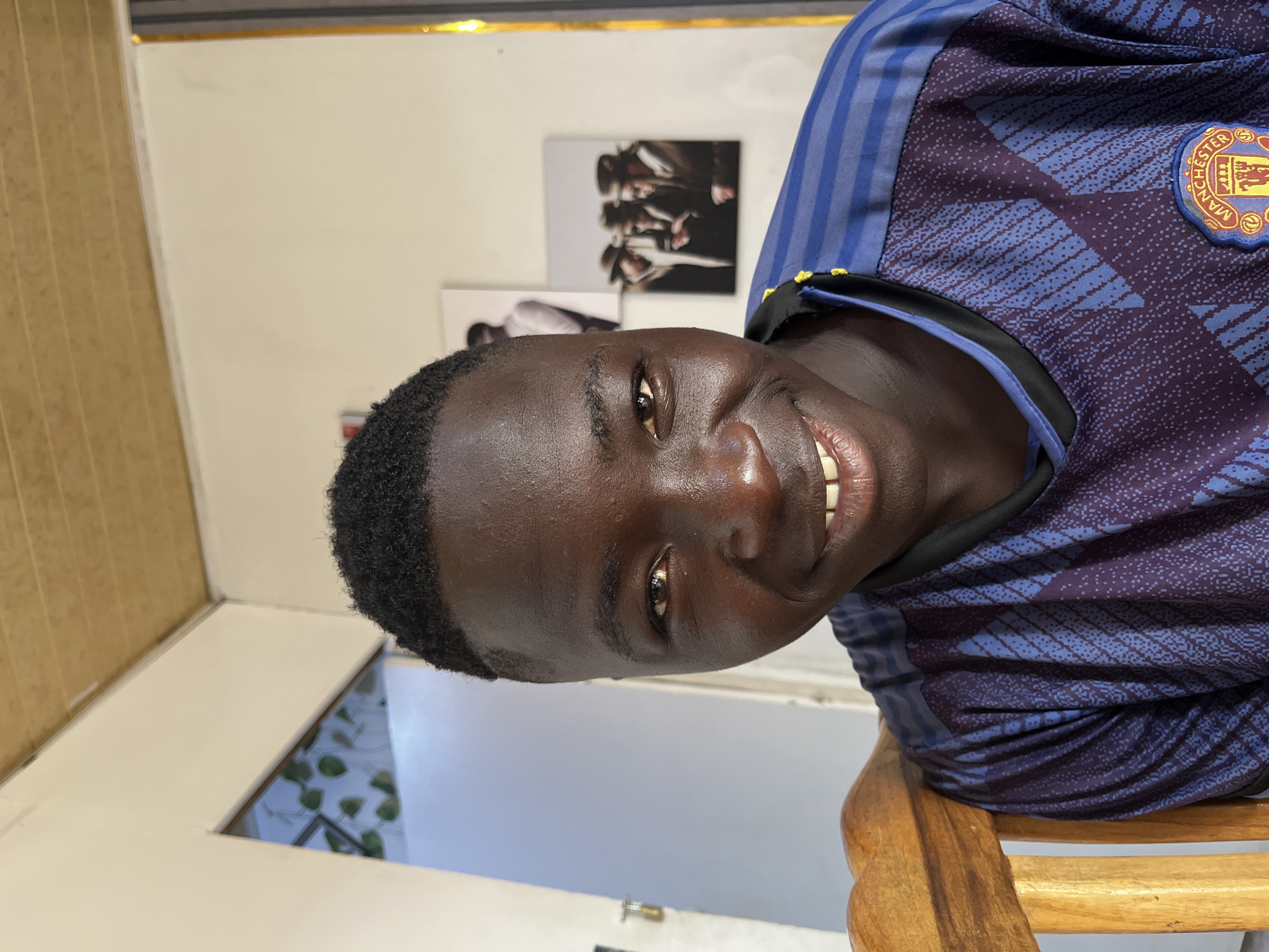A medium shot captures a smiling young man with dark skin and short, textured hair. He is wearing a blue Manchester United soccer jersey. He is seated on a wooden chair. Behind him are two framed pictures on a white wall. To the left, a section of a window with a view of greenery is visible. The environment suggests an indoor space, likely a home, in Jos, Nigeria.