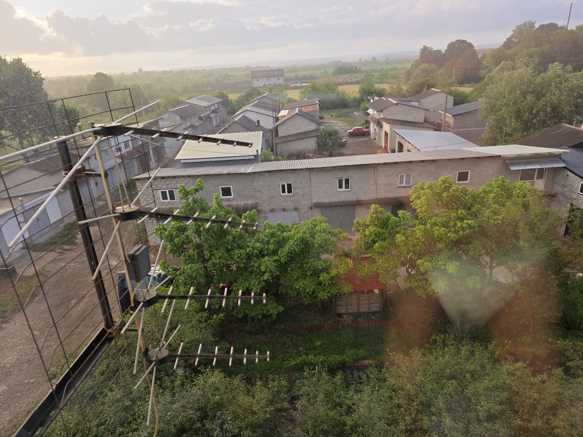 The image shows an aerial view of a rural residential area in Dubno, Ukraine. In the foreground, a television antenna is partially visible, obscuring parts of the scene. The scene depicts a collection of single-story and multi-story buildings, many with corrugated metal roofs. There are numerous trees and green vegetation throughout the area, indicating a lush environment. A red car is parked in the courtyard of one of the buildings. The sky is overcast with clouds, and the overall lighting suggests it might be early morning or late afternoon. The environment appears to be a well-established residential or light industrial area, likely a village or the outskirts of a town, given the density of buildings interspersed with greenery.