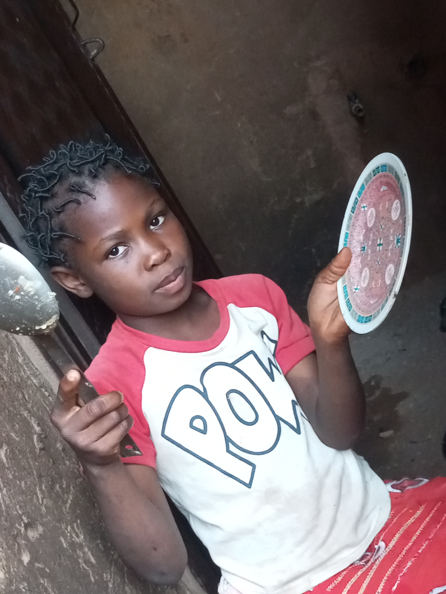 A young girl with braided hair stands in what appears to be a doorway in Tempure, Nigeria. She is wearing a red and white t-shirt with "POWER" written on it in large white letters, and a red skirt. In her left hand, she holds a metal spoon, while her right hand holds a decorative plate. The background is a rough, brown wall and a dark wooden door. The lighting is dim, suggesting it may be indoors or in a shaded outdoor area.