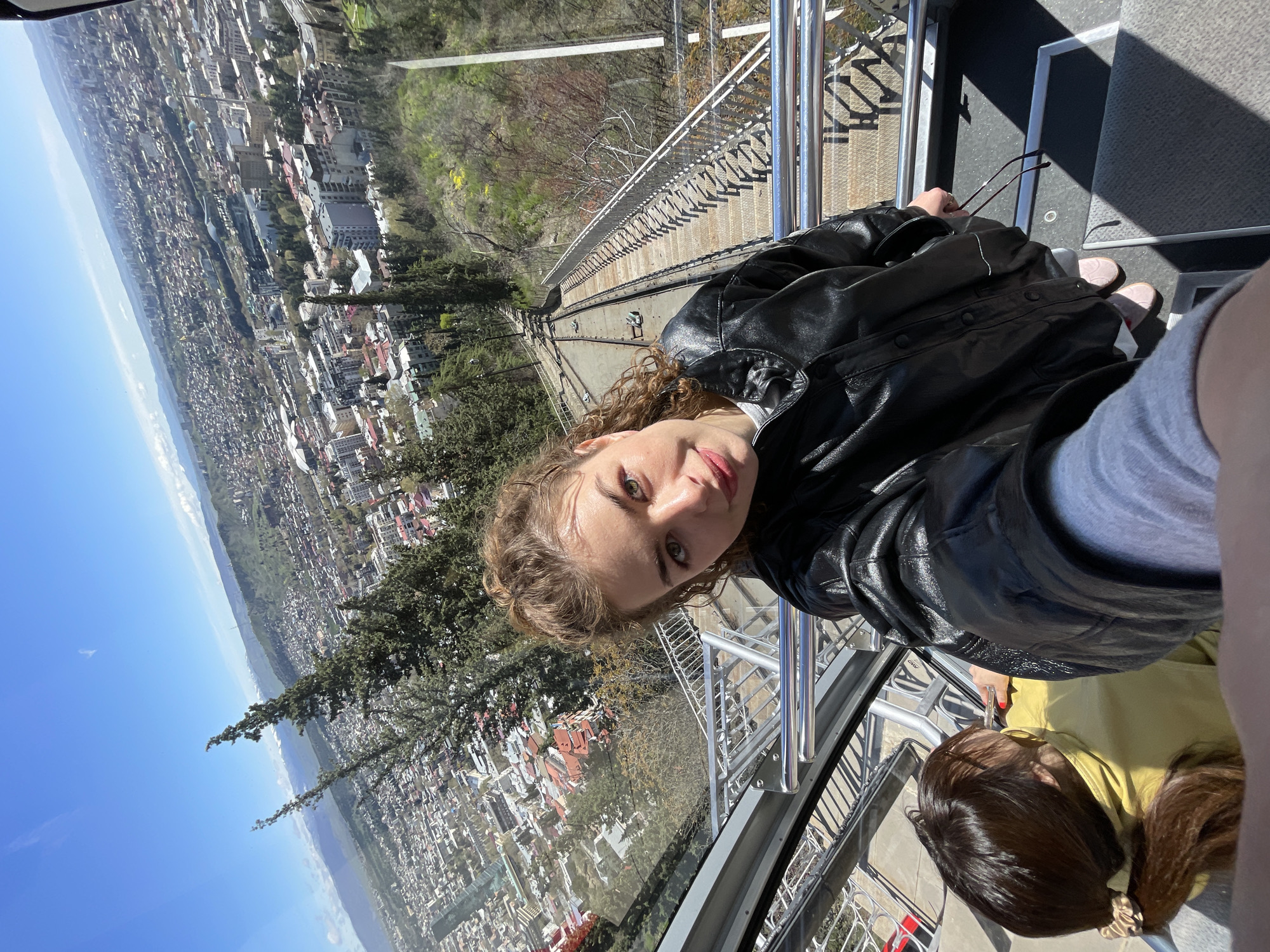 A rotated, eye-level shot features a woman in a black leather jacket looking at the camera. She appears to be on a funicular or cable car overlooking the city of Tbilisi, Georgia. The track of the funicular descends steeply into the cityscape below, which is visible through the metal railing and glass windows. The environment includes buildings, trees, and a blue sky. Another person with dark hair wearing a yellow shirt is visible in the lower part of the frame.