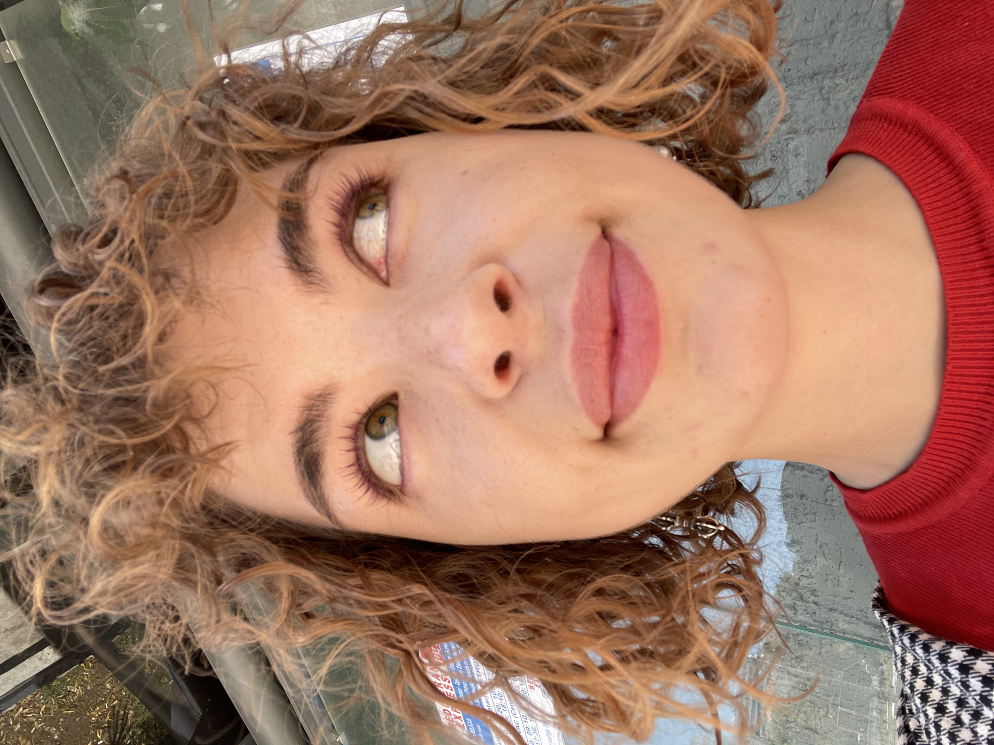 The image is a close-up, slightly tilted perspective shot of a young woman with curly, auburn-toned hair. She has fair skin, high cheekbones, and slightly parted, full lips adorned with pink lipstick. Her eyes are light green with makeup applied to her eyelids. She is wearing a red sweater. In the background, there is an out-of-focus view of Rafailovići, Montenegro, specifically displaying a window and reflections of the surroundings.