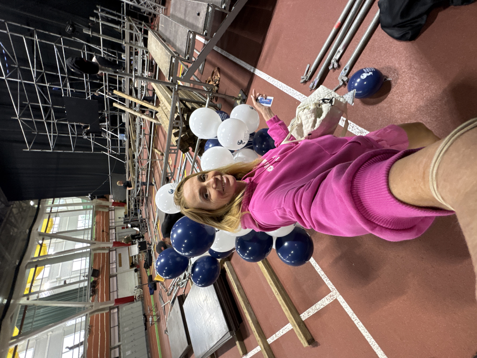 A woman with blond hair wearing a pink hoodie is lying on a reddish-brown floor with white lane markings. She is surrounded by a cluster of white and navy blue balloons. She is holding a small card in her hand. In the background, there's a large indoor space with a track visible, scaffolding structures, stage equipment, and window frames, indicating a large assembly area or indoor athletic facility. The location of Irinovka, Russia, is not discernible from the visual information provided.