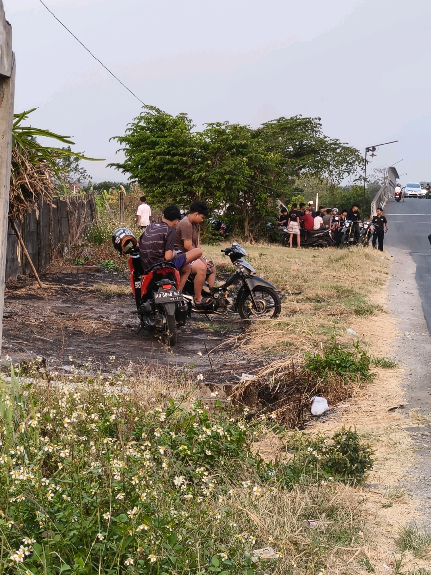 The image depicts a scene in Desa Sobokerto, Indonesia, with a focus on a group of young people gathered near motorcycles by the side of a road. In the foreground, two young men are seated on a red and black motorcycle. The motorcycle has a license plate reading "AD 6860 NZ". A helmet is placed on the back of this motorcycle. To the left, another black motorcycle is parked, partially disassembled, with its engine exposed. The ground around the motorcycles is dirt and dry grass, with some areas appearing scorched or burnt.
In the middle ground, a larger group of young people are gathered around several other motorcycles. They are standing and interacting, with some seated on the motorcycles. The environment includes dry vegetation, some scattered debris, and a wooden fence in the background. Further in the background, a road crosses over a bridge, with a few cars and motorcycles visible on the bridge. Trees are present on both sides of the road and bridge. The sky is overcast and appears to be late afternoon or early evening. The overall impression is of a casual gathering or break by a group of young individuals in a semi-rural setting.