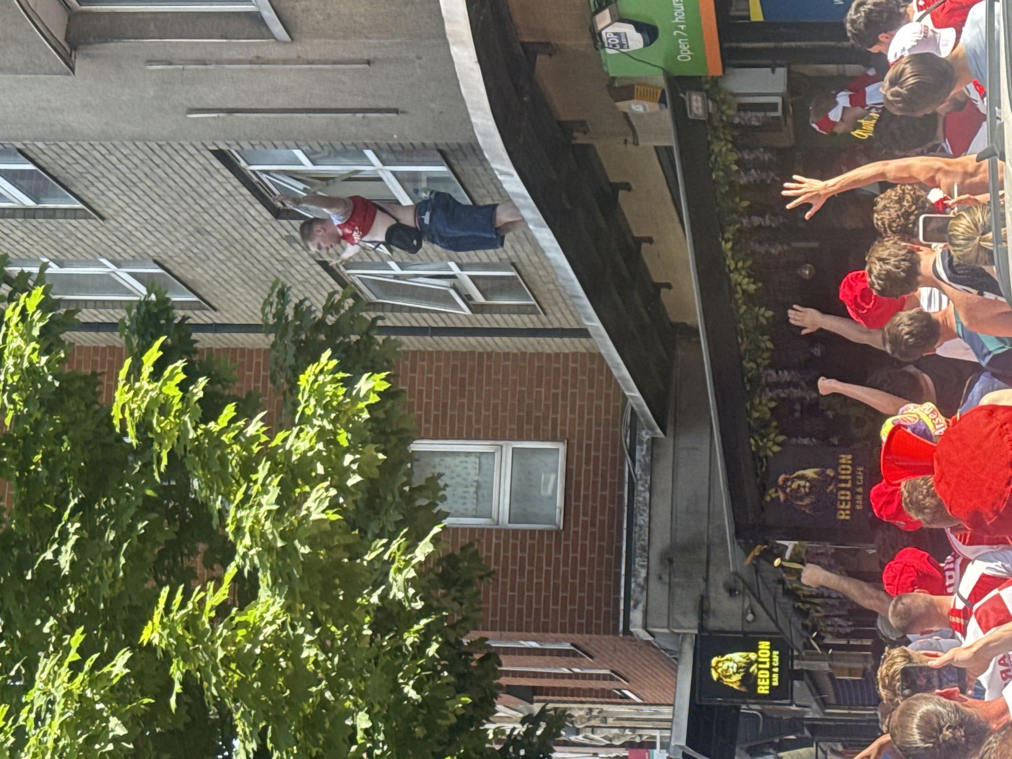 The image depicts a street scene in London, United Kingdom, likely during a sporting event celebration. A man wearing a red and white shirt and blue shorts is hanging out of a window of a multi-story brick building. He has a small black bag across his chest. Below him, on the street, is a large crowd of people, many of whom are wearing red clothing and raising their arms. A sign for "RED LION BAR & CAFE" is visible, suggesting the location is a public house. Other signs indicate the bar is open 24 hours and has a COP ALARME system. The environment includes foliage from a tree on the left and building facades in the background.