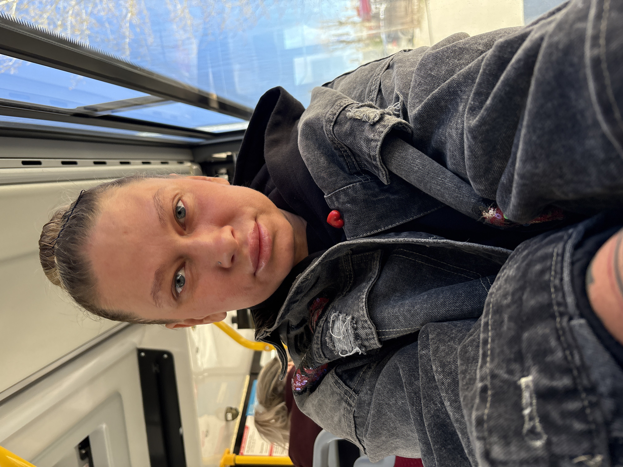 A woman with fair skin and blue eyes is seated inside what appears to be a public transport vehicle, possibly a bus or a tram. She is wearing a black hooded sweatshirt and a gray denim jacket. Her hair is pulled back. The vehicle has a large window to her left, through which a blurred cityscape is visible, potentially located in Perm, Russia. The vehicle's interior is light-colored with yellow handrails and panels.