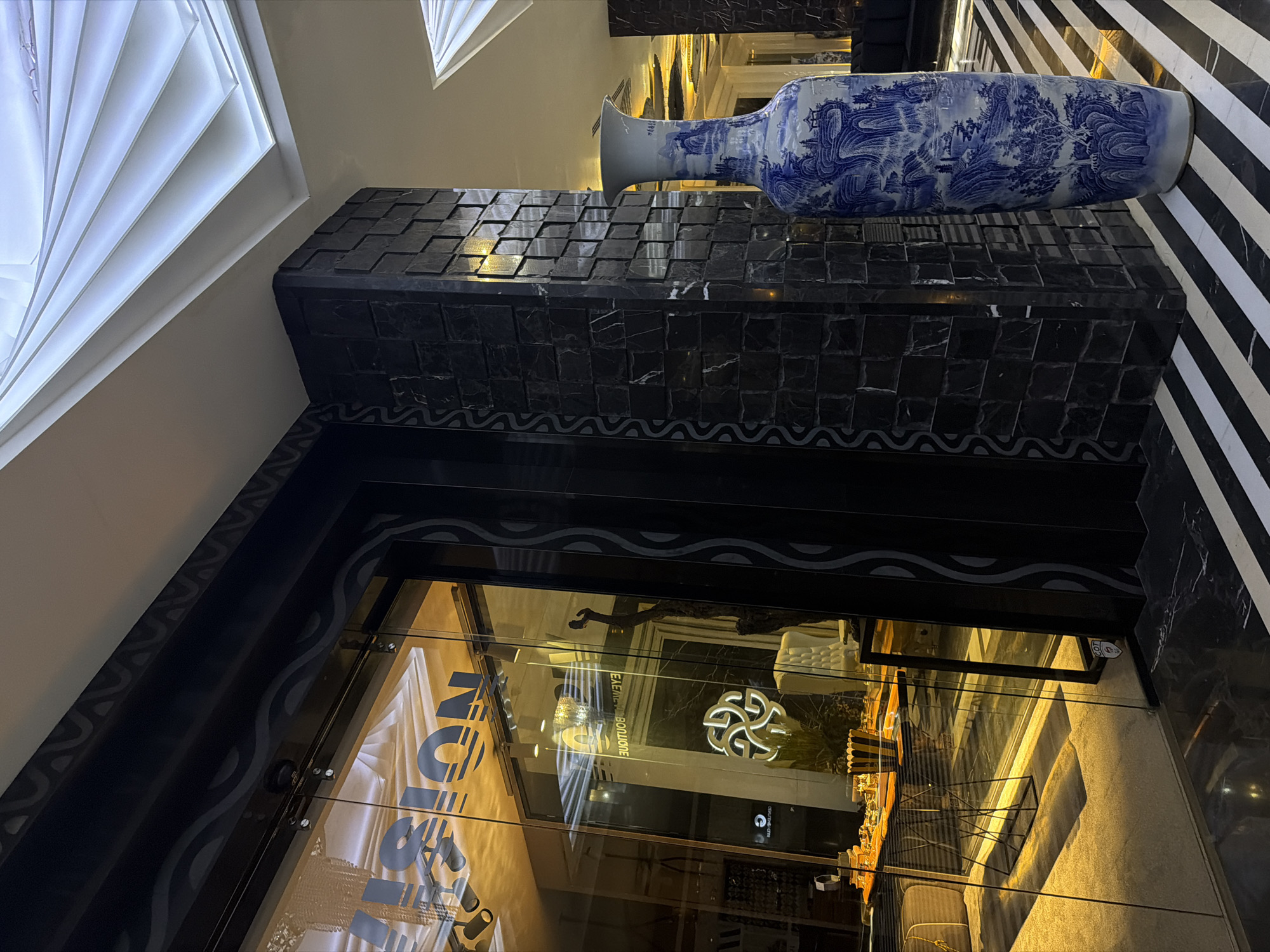 The image depicts the exterior of a retail establishment, specifically the "EXCESS BOUTIQUE," as reflected in the storefront window. The location is Sofia, Bulgaria. Above the store window sits a dark, possibly marble-tiled platform displaying a large, elongated porcelain vase decorated with blue and white scenic patterns. The building's exterior features a black and white striped design to the right of the vase. The interior reflection shows clothing and mannequins inside the store. The lighting in the store casts a warm, yellow glow. The angle and lighting conditions suggest the image was taken either early in the morning or in the evening.
