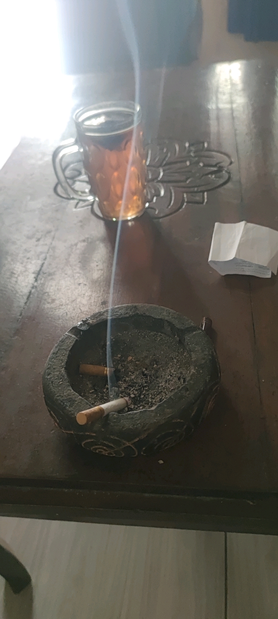 A glass mug of amber-colored tea sits on a patterned coaster on a wooden table. To the right of the mug, a small, folded piece of white paper rests on the table. In the foreground, a dark, circular ashtray contains ash and two cigarette butts. One cigarette butt is partially lit and emitting a thin stream of smoke that rises towards the mug. The ashtray appears to be made of stone or concrete and has a carved decorative pattern around its rim. The lighting in the image is bright, suggesting it was taken during the daytime, possibly indoors near a window. The overall scene suggests a casual, relaxed moment. The location is identified as Gambarsari, Indonesia.
