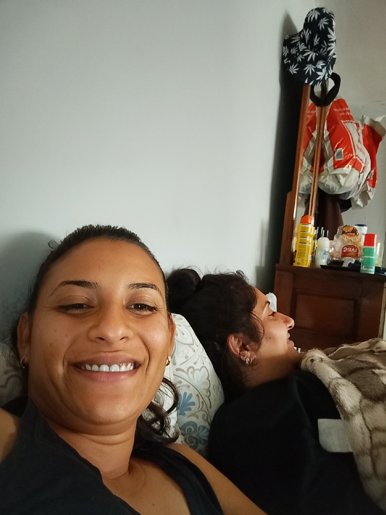 The image shows two women lying in bed. The woman in the foreground is facing the camera and smiling. She is wearing a black tank top. The woman in the background is lying on her side and appears to be asleep. She is wearing a black shirt and is covered by a blanket. There is a dresser with various items on it in the background, including bottles and a bag. A mirror is attached to the dresser. A hat with a leaf pattern is hanging on the wall above the dresser. The scene appears to be a bedroom.