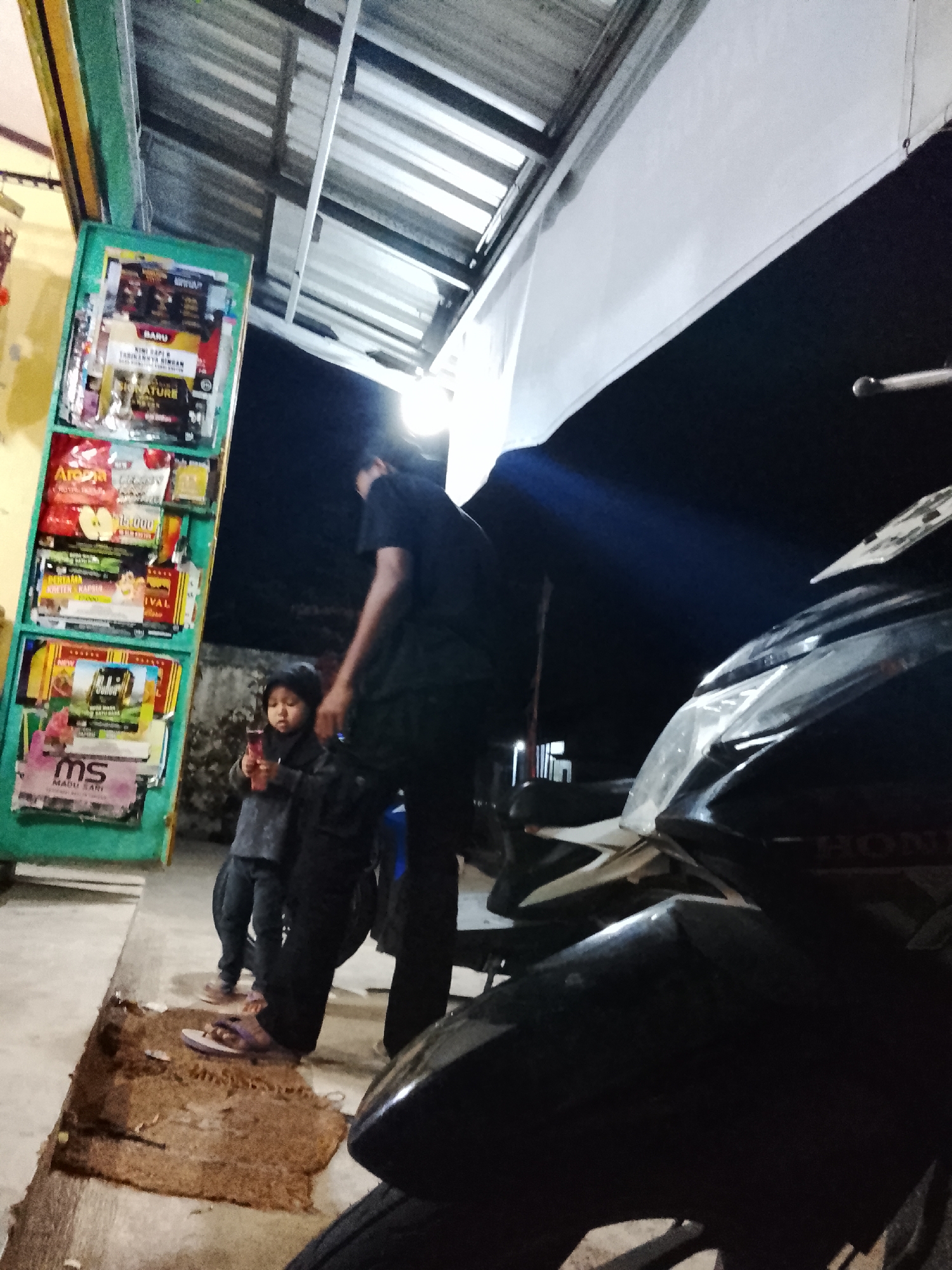 In Kalijaran, Indonesia, at night, a child stands holding a pink object in front of a display rack of magazines and other products. A person wearing a black t-shirt and pants stands behind the child, also facing the display. The display rack is green and filled with various items, including magazines with images of food and products. To the right of the scene, a black motorcycle is parked, its front visible with a license plate. The environment appears to be an outdoor area with a covered roof overhead. The ground is concrete, with a brown mat lying on it.