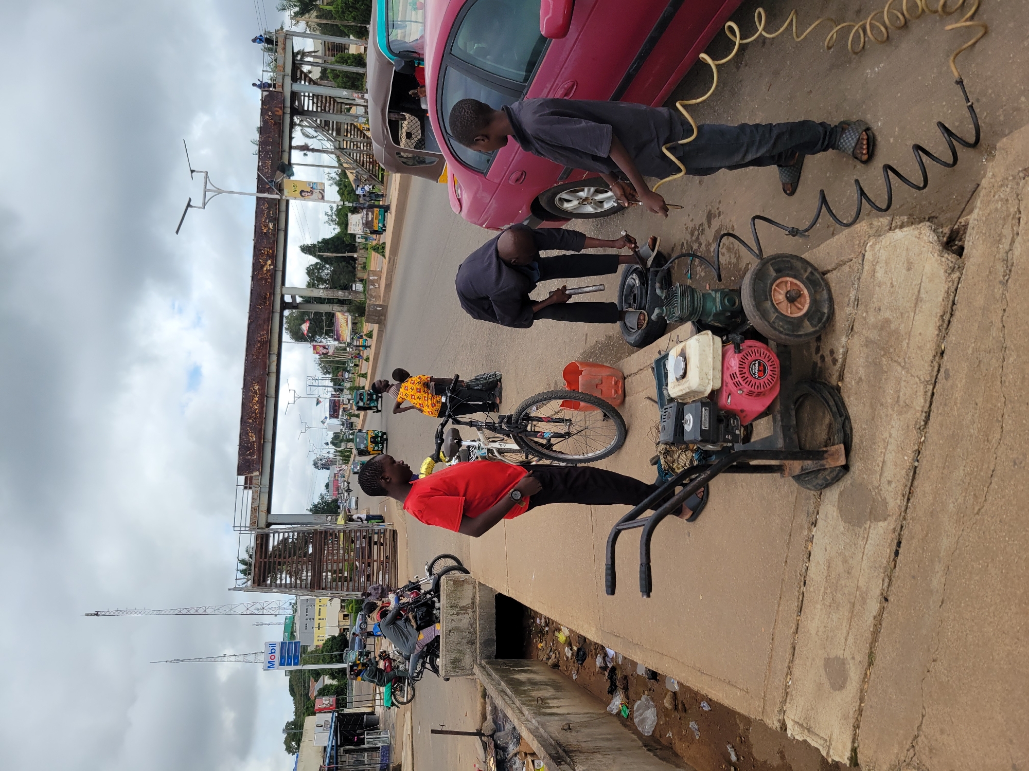 A group of individuals are engaged in roadside vehicle repair in Jos, Nigeria. The scene is set on a paved street with a concrete drainage ditch running alongside it, filled with debris. In the foreground, a red car is parked with its rear wheel off and resting on the ground next to an air compressor. Two young men are focused on the tire: one, wearing a dark shirt and jeans, appears to be attaching a hose to the wheel, while the other, in a black shirt and pants, is leaning over and working on the tire itself. To the left, a man in a red shirt stands near a bicycle and an air compressor, observing the activity. Further down the street, several motorcycles and a yellow auto-rickshaw are visible, along with a pedestrian bridge overhead. The sky is overcast.