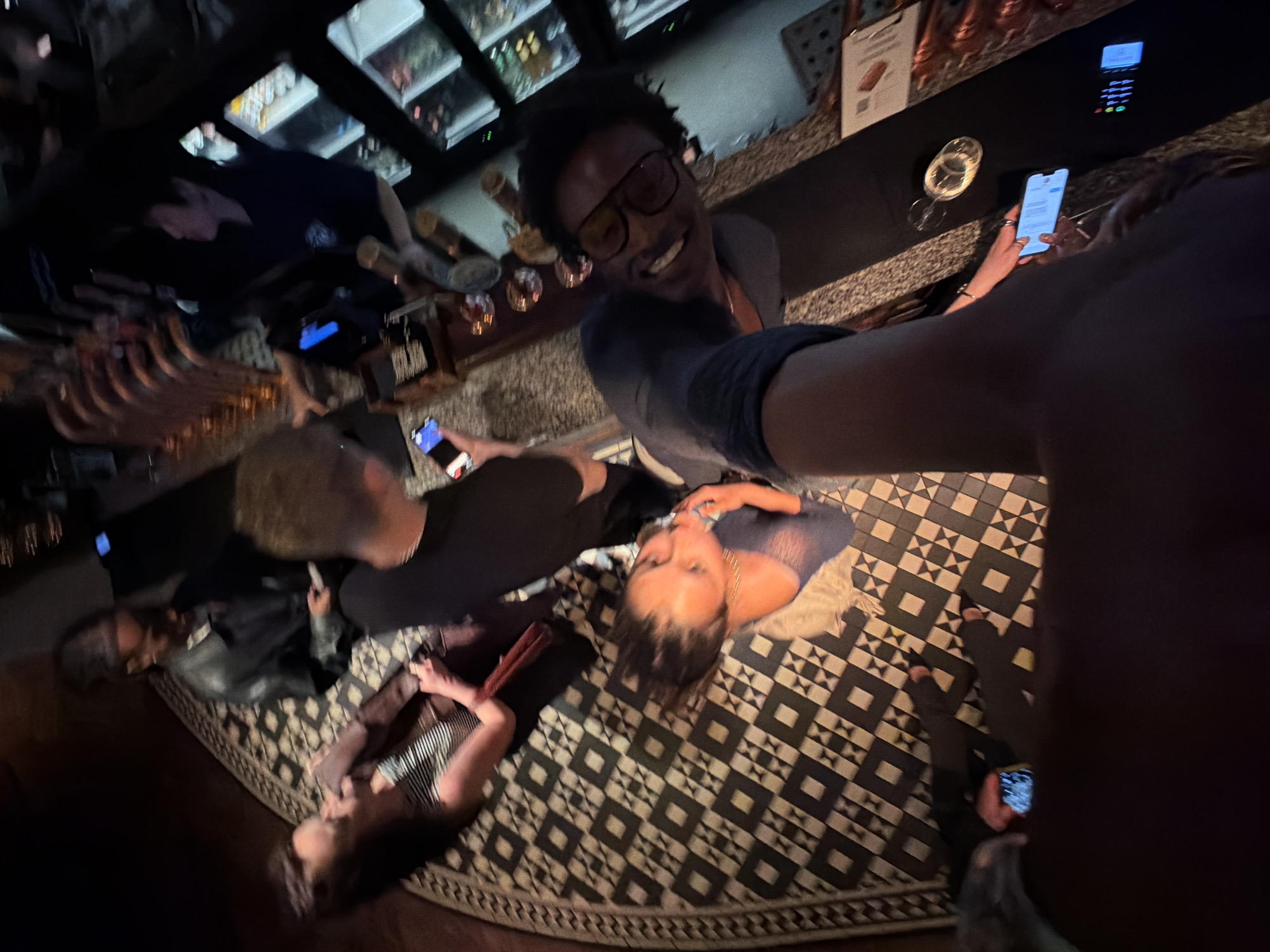 This image captures a vibrant, dimly lit scene within what appears to be a bar or lounge in London. The perspective is a high-angle, wide-lens shot, possibly a selfie, creating a dynamic view of a social gathering.
In the center-right, a man with dark skin, dark curly hair, and glasses, wearing a dark shirt, smiles directly at the camera. His arm extends towards the right, suggesting he might be holding the camera. To his lower left, a woman with dark hair and a light-colored top (possibly mesh) is reclining or lying on a large, patterned rug, looking up towards the camera.
Several other individuals are present. One person with short, light brown curly hair and a dark shirt is standing or sitting further back, looking down at a mobile phone. Another person in a black jacket is visible on the far left. On the bottom left, a person is partially visible, also on the rug. Multiple hands holding illuminated mobile phones are seen throughout the image.
The background features a bar counter with visible bottles on shelves behind it, characteristic of a nightlife venue. A payment terminal and a glass containing a drink are also on the counter. A sign displaying a QR code is visible on the bar counter. The floor is dominated by a large, circular rug featuring a striking black and white geometric pattern of squares and crosses.
The overall atmosphere is social and relaxed, indicative of an evening or nighttime gathering. The dim, warm artificial lighting reinforces the time of day.