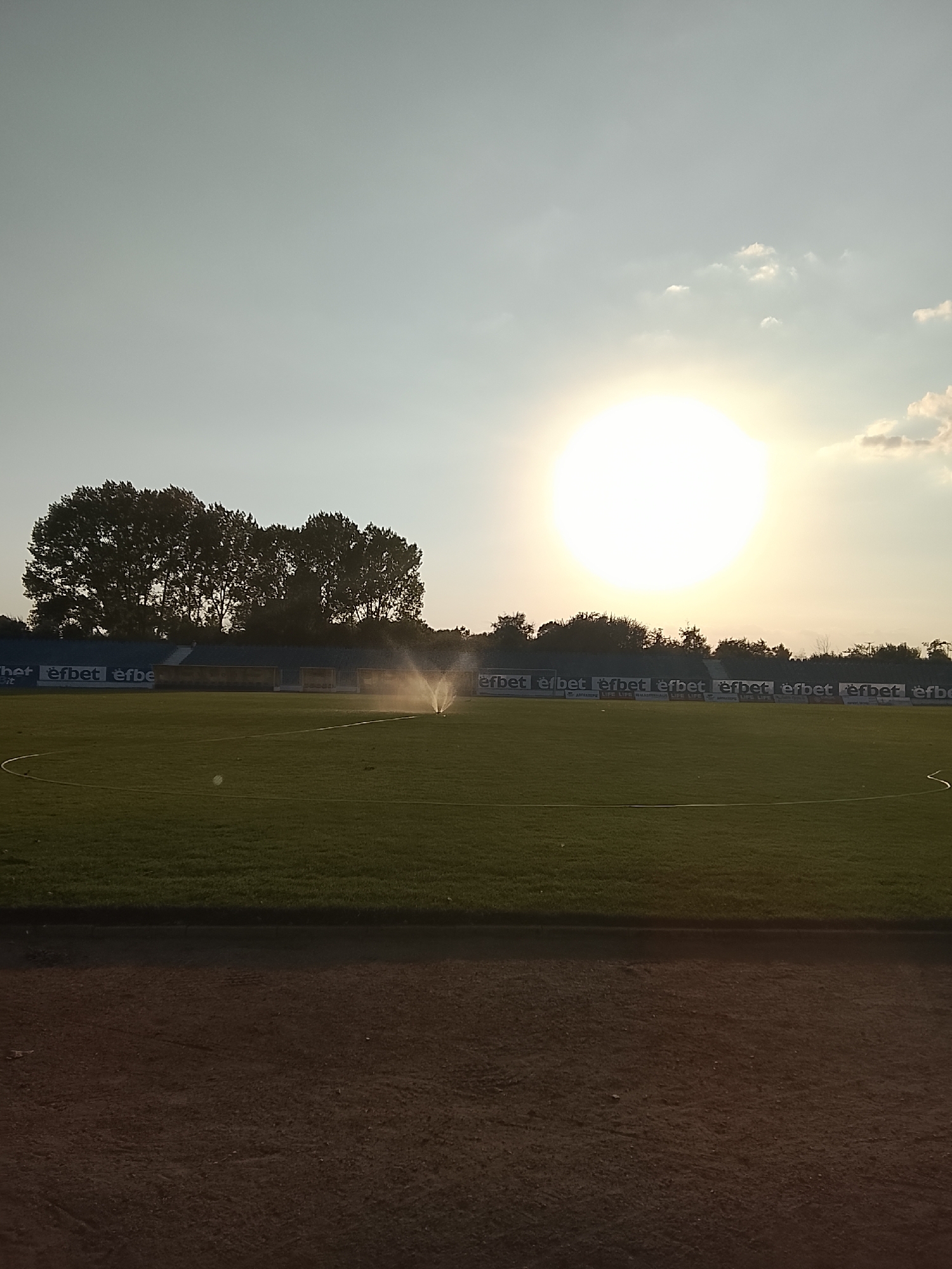 The image captures a wide shot of a sun-drenched soccer field. The sun is prominently featured, a large, bright orb in the upper right quadrant of the sky, casting a warm glow. In the background, a line of tall, leafy trees stands silhouetted against the sky. Below the trees, a stadium grandstand is visible, with blue seating and advertising banners along the front, displaying "efbet" repeatedly. The green grass of the soccer field fills the middle ground, and a sprinkler system is actively watering the field, creating a misty spray. In the foreground, a dirt track or path runs along the bottom of the image. The overall scene suggests an outdoor sporting venue, possibly during a late afternoon or early evening.