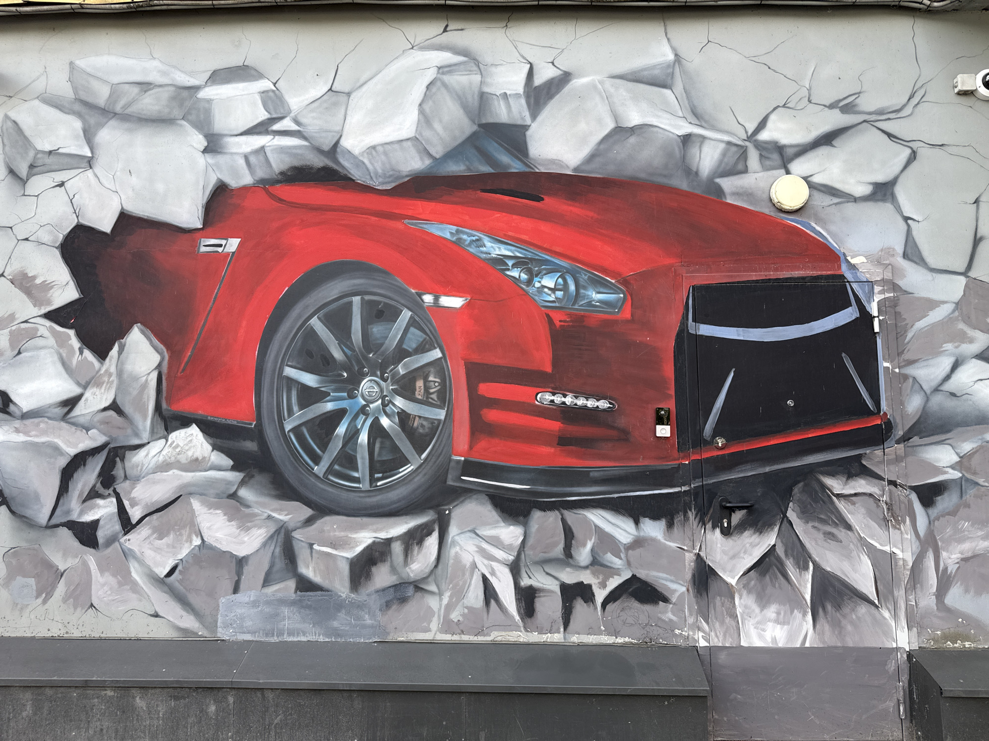 An exterior wall in Saint Petersburg, Russia, features a detailed mural depicting a red Nissan GT-R sports car. The artwork creates a trompe l'oeil effect, showing the car emerging from a shattered gray wall, surrounded by painted white and gray debris and dark cracks. The visible portion of the car includes its front-left quarter, highlighting a silver multi-spoke wheel with a visible Nissan logo and a brake caliper, detailed headlights, a section of the hood, and a black front bumper/grille. A silver "GT-R" emblem is painted on the vehicle's side fender. Real-world fixtures are integrated into the painted scene: a black utility panel with a handle on the lower right is painted to blend with the car's bumper and the surrounding wall debris. A circular white ventilation cap is mounted on the wall above the car's right front fender. A white security camera is positioned on the upper right section of the wall. Electrical conduits run horizontally along the top edge of the wall. A dark gray base structure is present below the mural. A small white rectangular tag is affixed to the painted car's right fender, and another small black rectangular object is on the lower portion of the car's body. No individuals are present in the image.