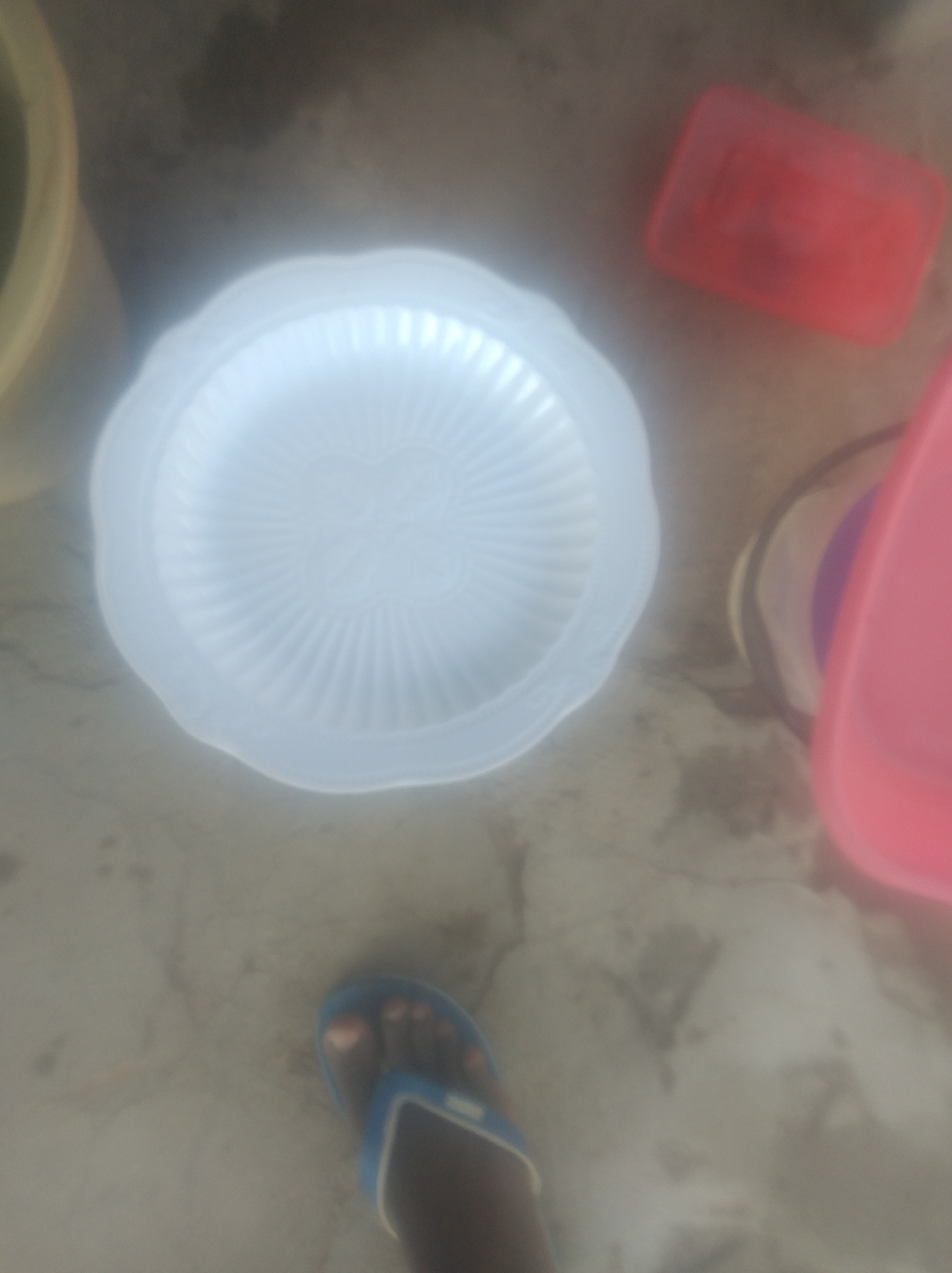 The image depicts a top-down perspective of a floor, likely outdoors, in Jalingo, Nigeria. Several objects are scattered across the concrete surface. On the left, a portion of a beige bucket is visible. To its right, a translucent white plate with a ribbed pattern is centrally placed. Further to the right, a bright red rectangular container is positioned. In the upper right quadrant, a portion of a pink object, possibly another container or lid, is visible. In the lower portion of the image, a person's foot wearing a blue flip-flop is visible, indicating a casual setting. The ground appears to be a rough, weathered concrete surface with some discoloration and debris. The overall impression is of an outdoor area, possibly a courtyard or yard, where items have been left or are in use.