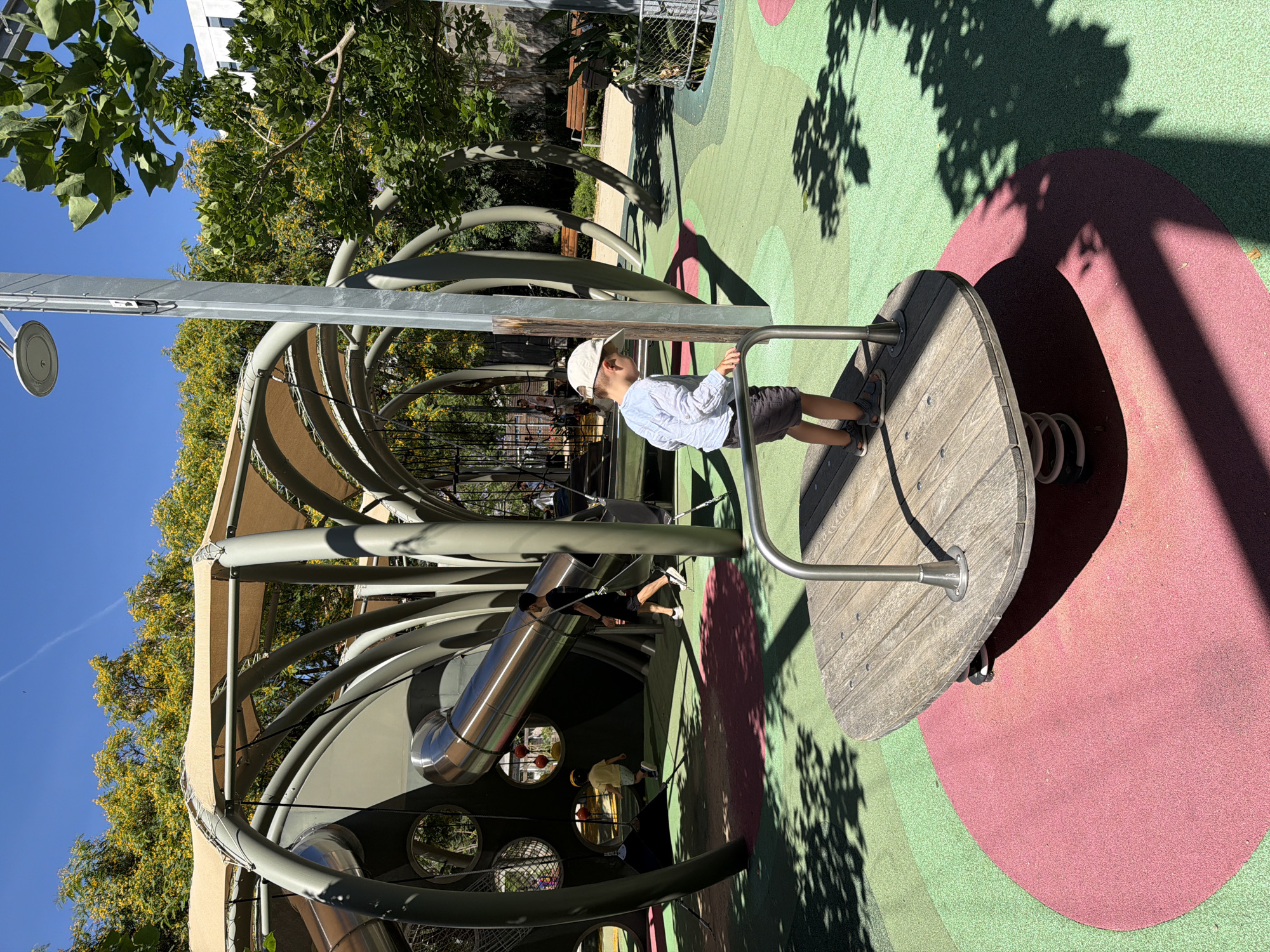 A young boy wearing a white baseball cap, light blue long-sleeved shirt, and dark shorts stands on a round, wooden spinning platform at a playground. He is holding onto a metal bar. The platform is supported by springs and is situated on a colorful, padded playground surface with red and green circular sections. In the background, a large, modern play structure with metallic tubes and beige fabric awnings is visible. A silver slide curves downwards from this structure. Other children and adults are present in the background, with some walking and others playing. Trees with yellow flowers and green leaves surround the playground, and the sky is clear and blue. This scene is located at the Parc del Mecat de Sant Pere, in Barcelona, Spain.