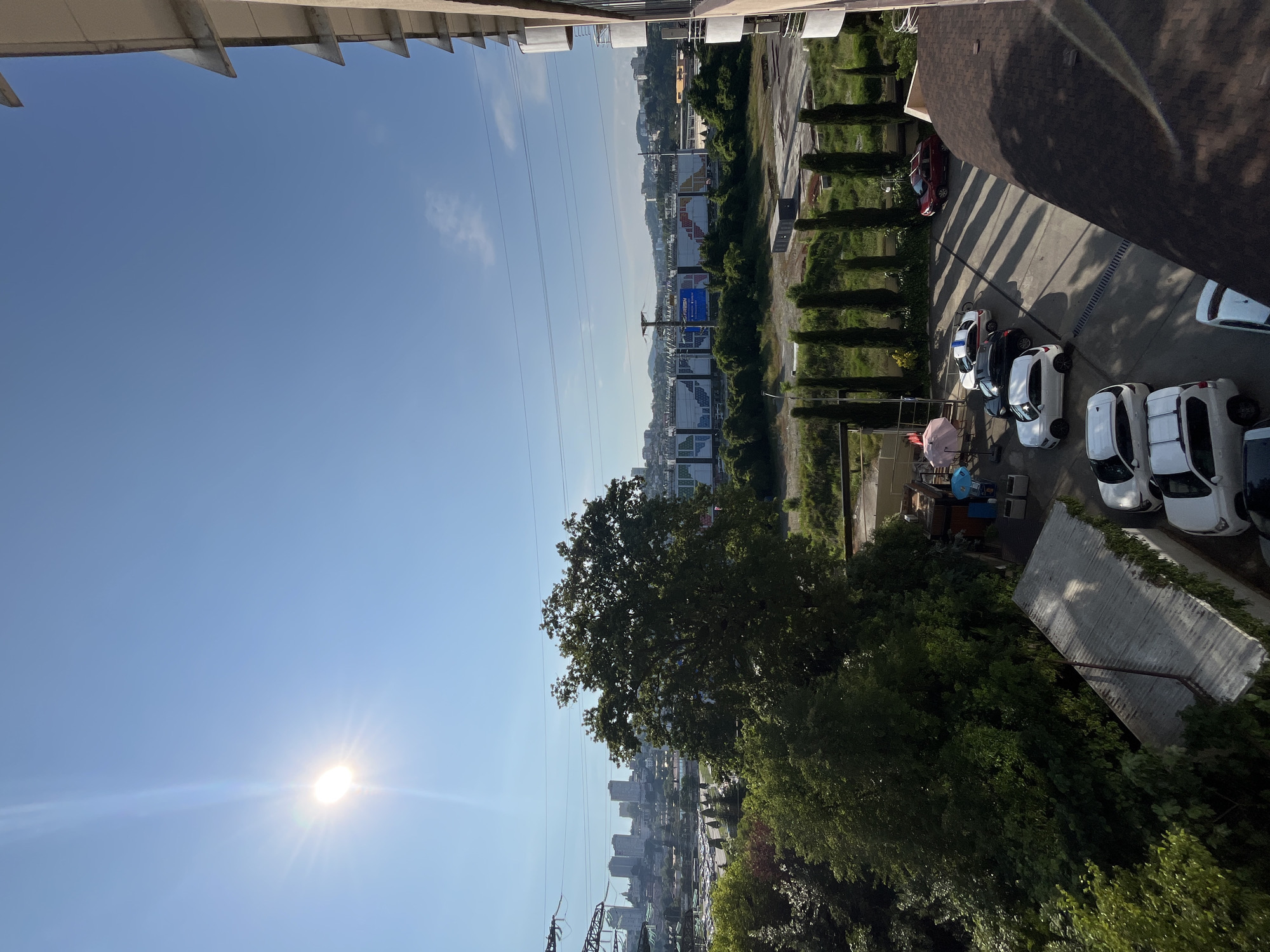 The image depicts a bright, sunny day in Sochi, Russia, with the sun shining prominently in the clear blue sky. The scene overlooks a residential building's balcony, offering a view of a street lined with parked cars, lush green trees, and distant city buildings. In the background, a large stadium or event venue with multiple banners and structures is visible. The overall impression is one of urban development integrated with nature, characteristic of a coastal city.