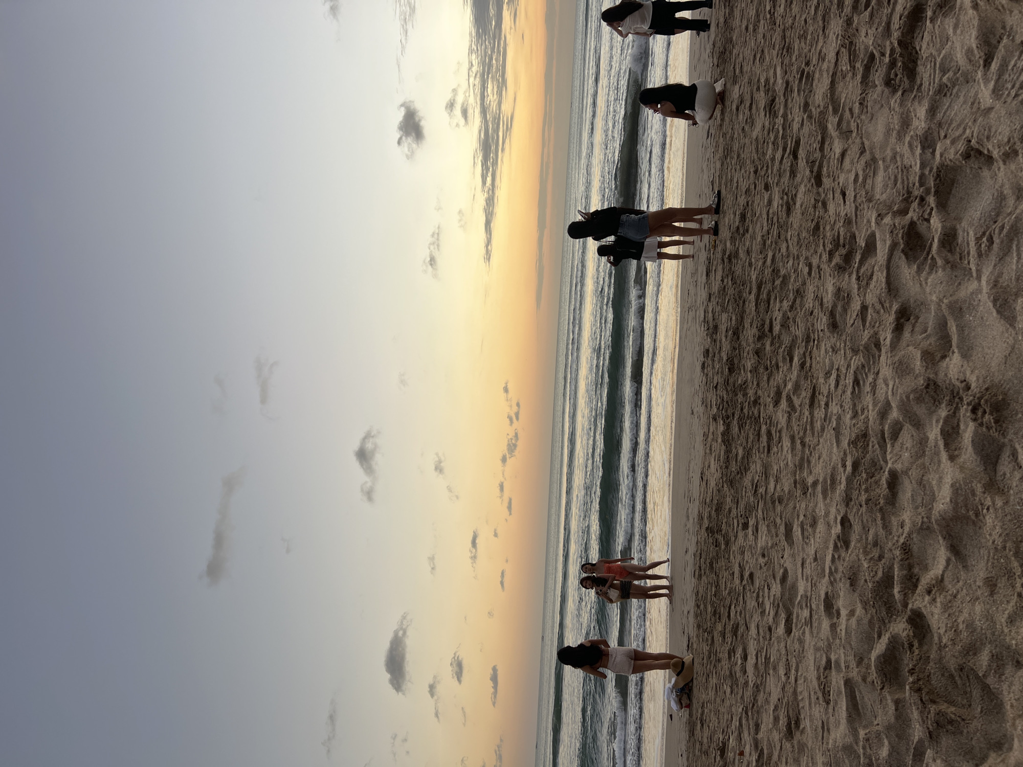 This image captures a serene beach scene at sunset in Kuta, Indonesia. The sky displays a gradient of soft blues and warm oranges, with scattered clouds adding texture. The ocean stretches out to the horizon, with gentle waves lapping at the shore. A sandy beach occupies the foreground, with footprints and ripples indicating recent activity.
Several individuals are visible on the beach, scattered at varying distances from the water. On the left side, three individuals are standing near the water's edge, two of whom appear to be talking. A fourth person stands further back, facing the ocean. In the middle ground, a group of three people are positioned, with one person appearing to hold up a phone, possibly taking a photo. Towards the right side of the frame, two individuals are visible, one kneeling and the other standing. A straw hat and some clothing items are placed on the sand near the left edge of the image. The overall atmosphere is one of peaceful observation of the natural beauty.