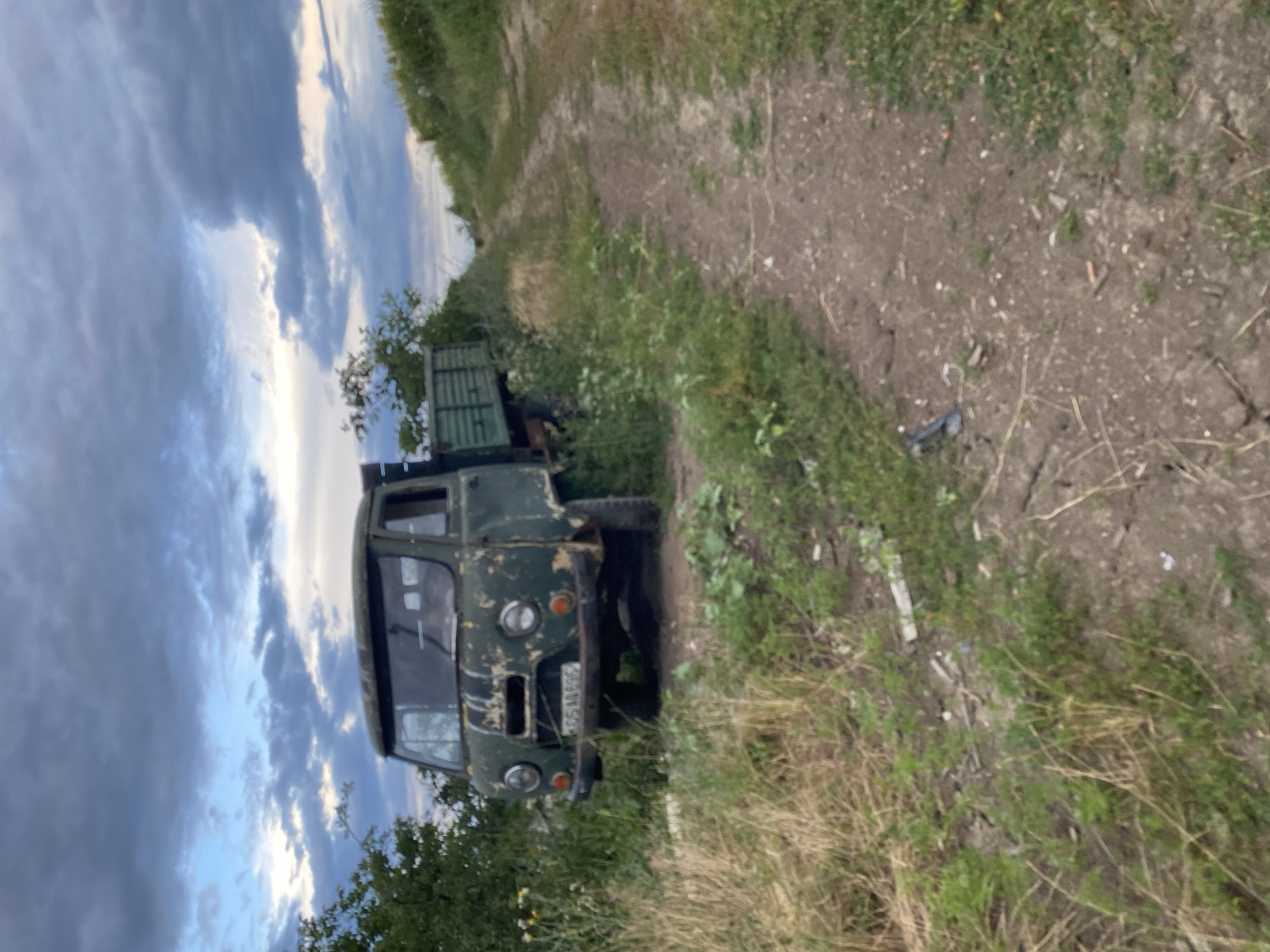 An old, weathered, green truck is parked on the side of a dirt road in Horodca, Moldova. The truck is partially obscured by tall grass and weeds. The road appears to be unpaved and is lined with vegetation on both sides. In the background, there is a cloudy sky. The scene suggests a rural or undeveloped area.