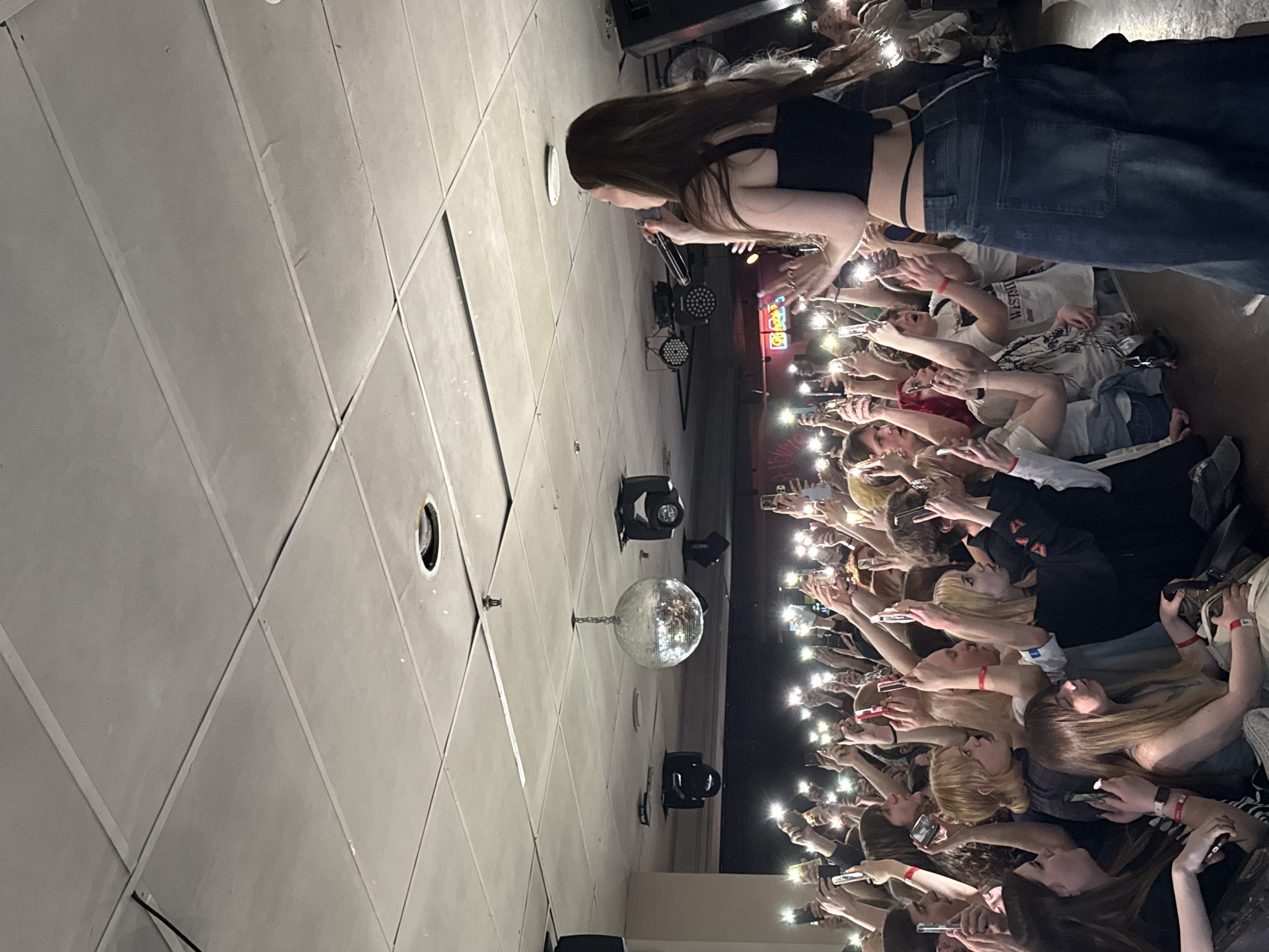 The image shows a concert or performance, possibly indoors at a venue in Krasnoyarsk, Russia. A female performer is on stage, holding a microphone and singing to a crowd. The audience is holding up their cell phones, using the flashlights, creating a bright light display in front of the stage. The performer is wearing a dark top and dark-wash jeans. The ceiling of the venue is visible and features a mirrored disco ball and stage lighting equipment. The audience is a diverse group of people with varying hair colors.