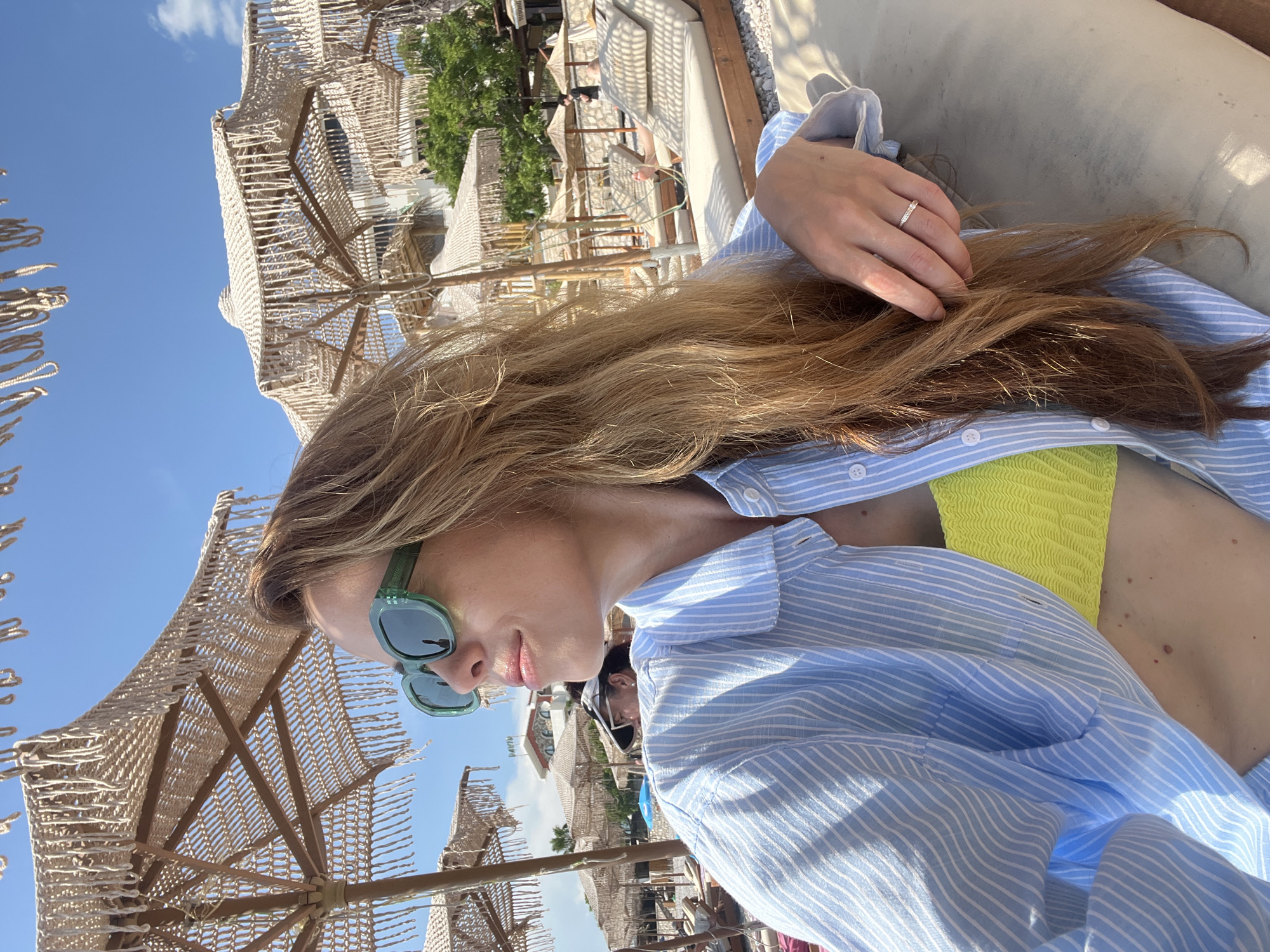 A blonde woman with long, auburn hair and green sunglasses is lying on a beach lounger on a sunny day. She is wearing a light blue and white striped unbuttoned shirt over a bright yellow bikini top. Her right hand rests on her lap, and she wears a silver ring with small diamonds on her ring finger. In the background, several thatched beach umbrellas with fringe detailing are visible, along with more beach loungers and some buildings and trees. The scene is set in Dobra Voda, Montenegro, a coastal town known for its beaches and clear waters. The sky is a bright blue with a few white clouds.