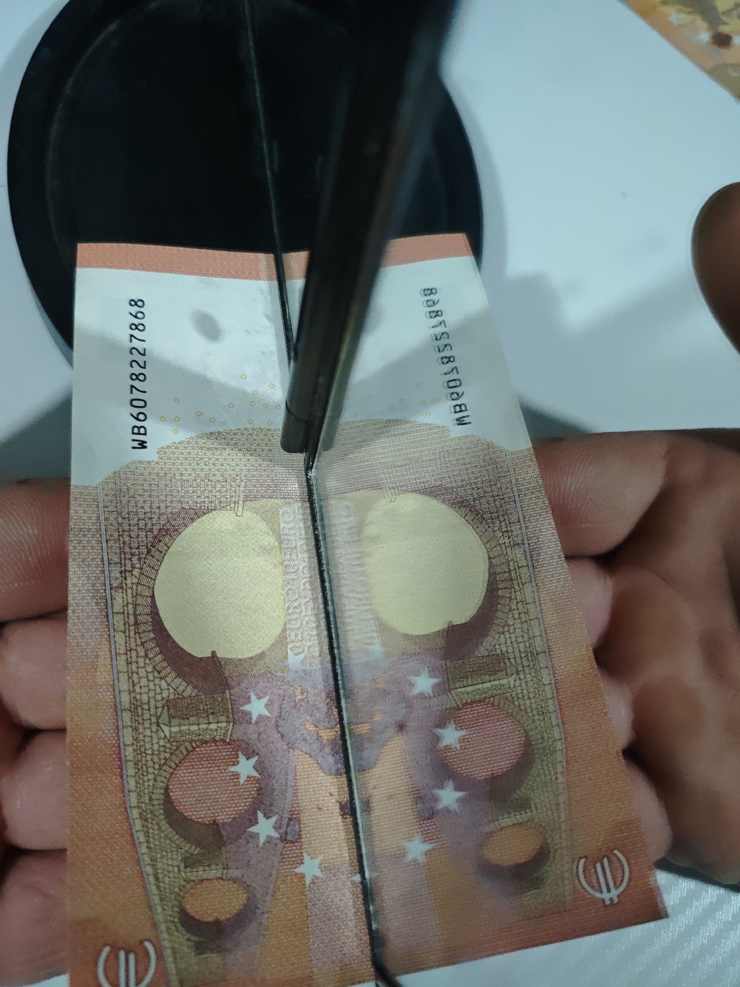 A pair of hands is shown holding a 50 Euro banknote, which is split vertically in the center by a dark, thin object, possibly part of a money-checking device or display mechanism. The banknote appears to be held over a light-colored surface, with a dark, circular object visible in the background, possibly part of the same device. The split allows both halves of the banknote to be visible, revealing security features like the transparent window with holographic elements and part of the watermark. The note is an orange-brown color with stars and the Euro symbol visible.
Visible text on the left side of the banknote includes the serial number "WB60782227868". On the right side, a partial serial number "MB001855389" is visible. The Euro symbol (Є) is present on the bottom right corner of the note. Parts of the word "EYPΩ EYPO" and "EURO" can be seen within the transparent window design. The number "50" is also incorporated into the note's design. The overall impression is that the banknote is being examined or displayed, possibly to highlight its security features or demonstrate a specific handling method.