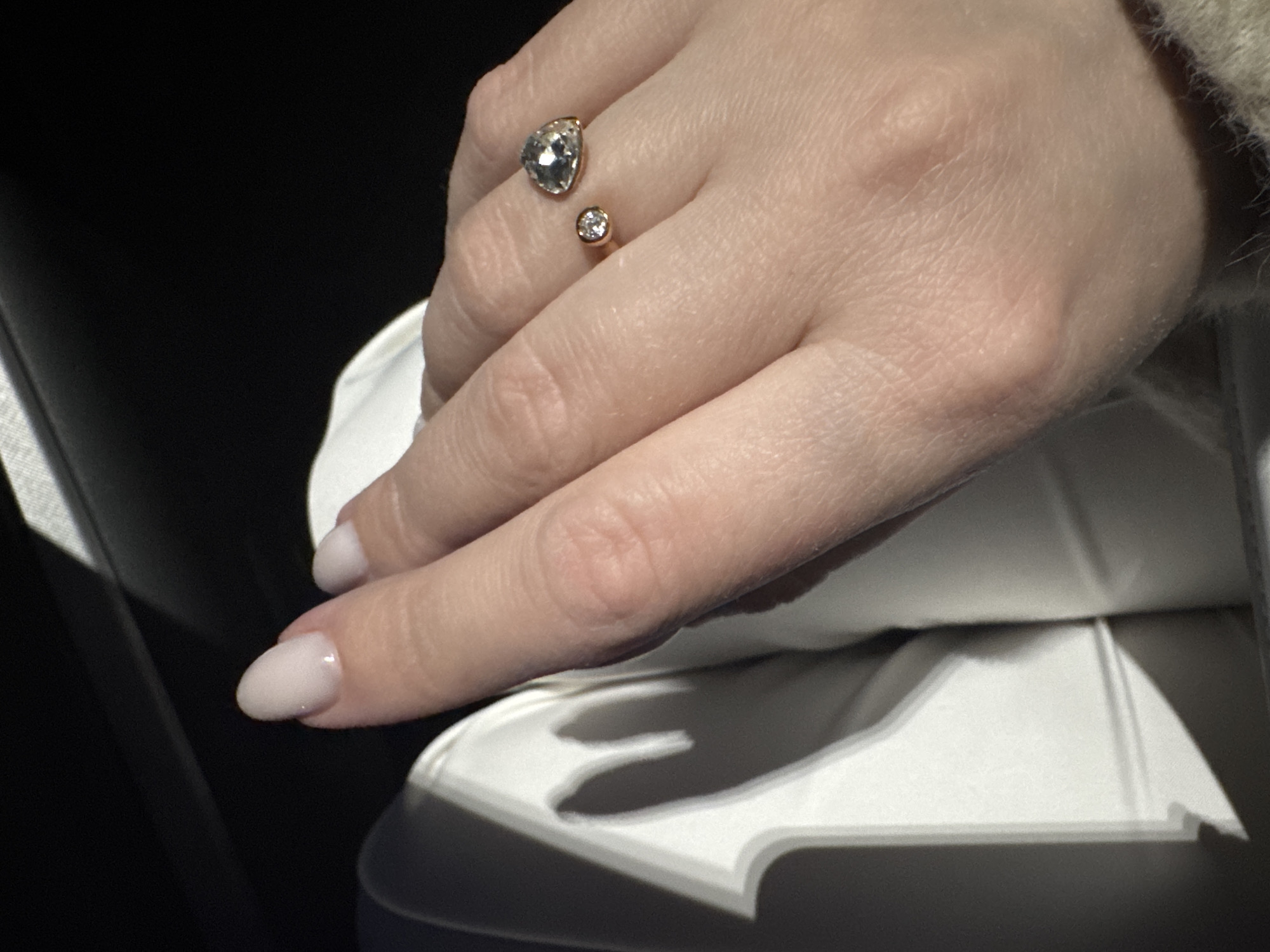 A close-up shot captures a person's hand adorned with a two-stone ring. The ring, appearing to be gold, features a larger pear-shaped stone and a smaller round stone. The hand rests on a white surface with defined shadows, suggesting an interior setting. The fingernails are painted a light color. The presence of shadows and the absence of specific landmarks prevent location determination in Chaikovsky, Russia, or any other location. The image's focus is on the hand and ring.