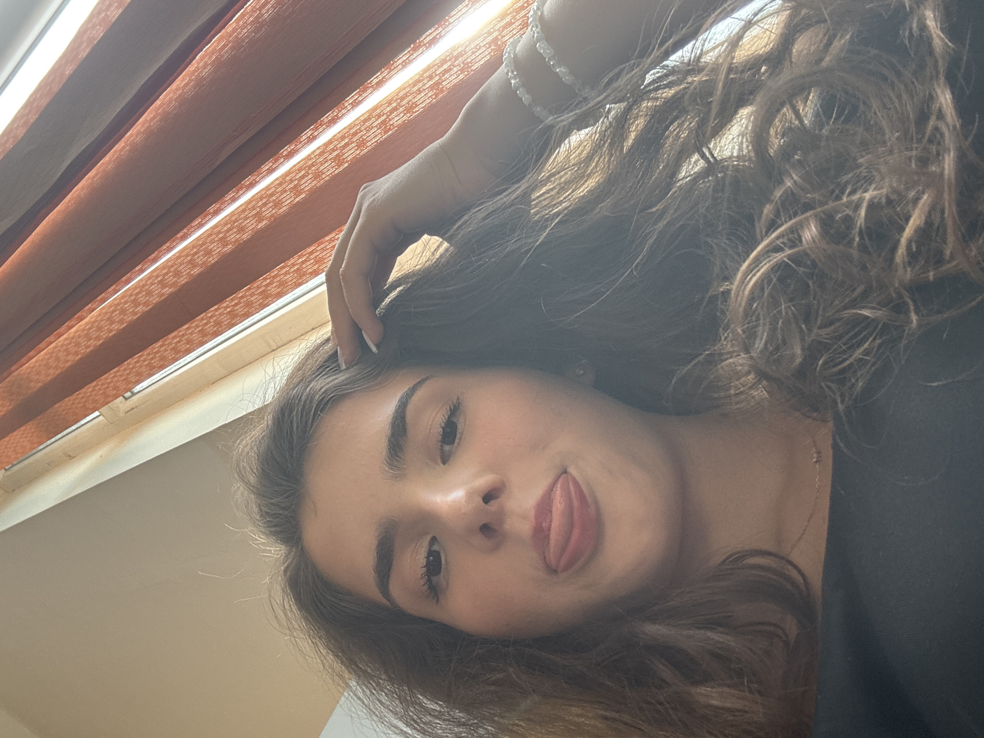 A young woman with long, wavy brown hair is captured in a close-up, low-angle selfie. She is sticking her tongue out slightly, with her head tilted. She is wearing a black top and a delicate gold necklace. On her left wrist, she wears two thin, beaded bracelets. Her right hand is resting on her head, with her fingers splayed. The background shows vertical orange blinds, suggesting the image was taken indoors, possibly near a window. The lighting is natural, coming from the side, casting soft shadows. The image does not provide any specific clues about its location within the city of Shumen, Bulgaria.
