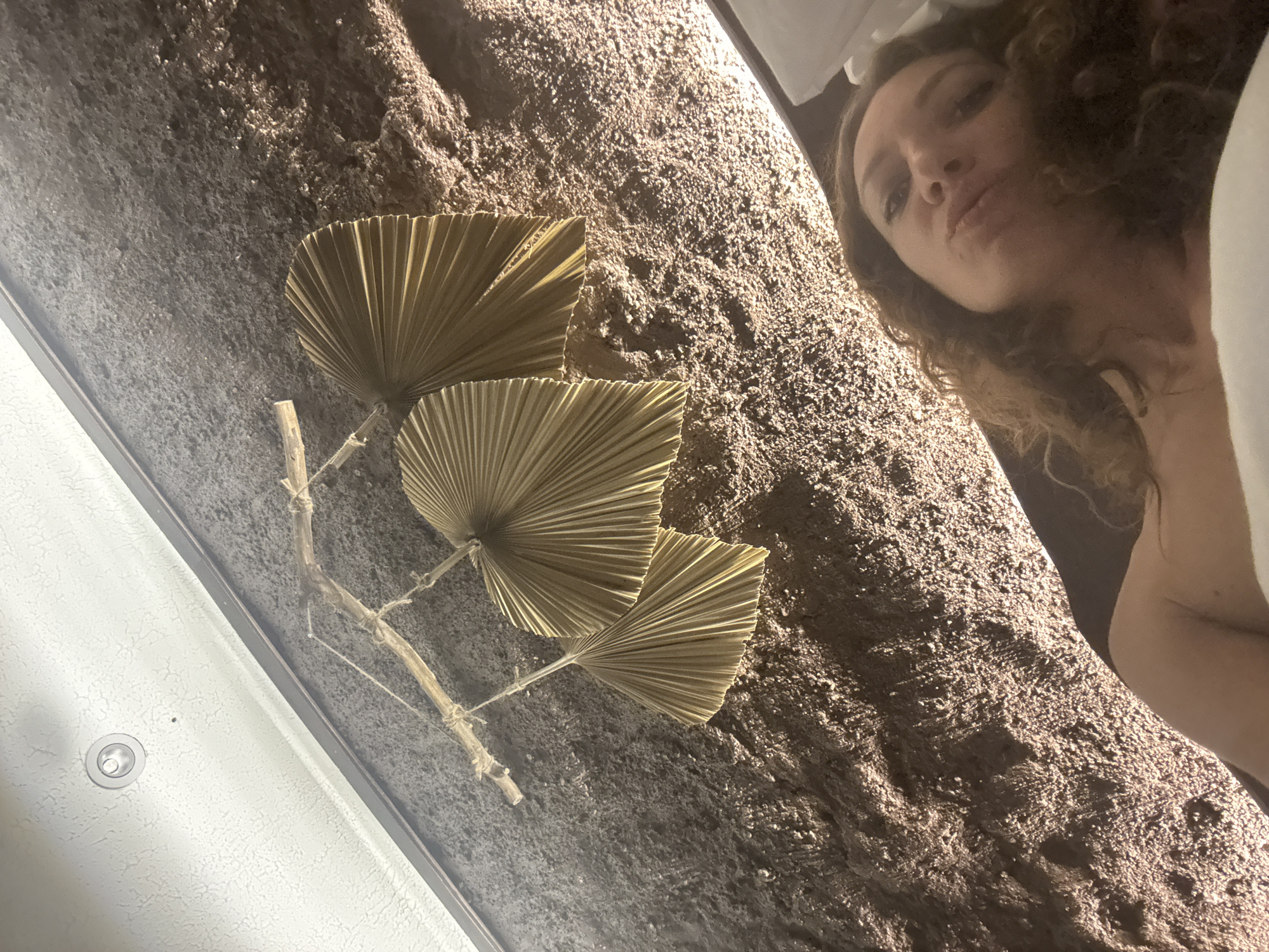 In Nam Hoa Lư Ward, Vietnam, a low-angle shot captures a woman lying down, her face tilted towards the camera in a pout. She is partially covered by a white sheet or towel. Above her, a rustic decoration made of dried palm leaves hangs from a twig, suspended by twine. The wall behind her has a textured, stone-like appearance. The ceiling, visible in the upper left, is white with a recessed light fixture. The overall lighting suggests an indoor, possibly dimly lit environment.