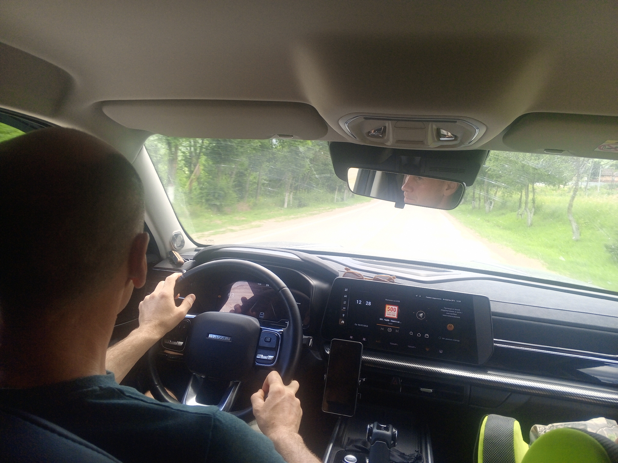 The image shows the interior of a car, looking forward from the passenger's perspective, with the driver's hands on the steering wheel. The driver, a man with short hair, is wearing a dark t-shirt. The car's dashboard is visible, featuring a large touchscreen display showing music information and navigation. A smartphone is mounted on the dashboard. Outside the car, a road is visible, lined with trees and greenery, suggesting a rural or semi-rural setting. The reflection in the rearview mirror shows another person, presumably the driver, with short dark hair and fair skin, looking forward. The image is taken in daylight. There is no visual information that can confirm the location as Голубинское сельское поселение, Ukraine.