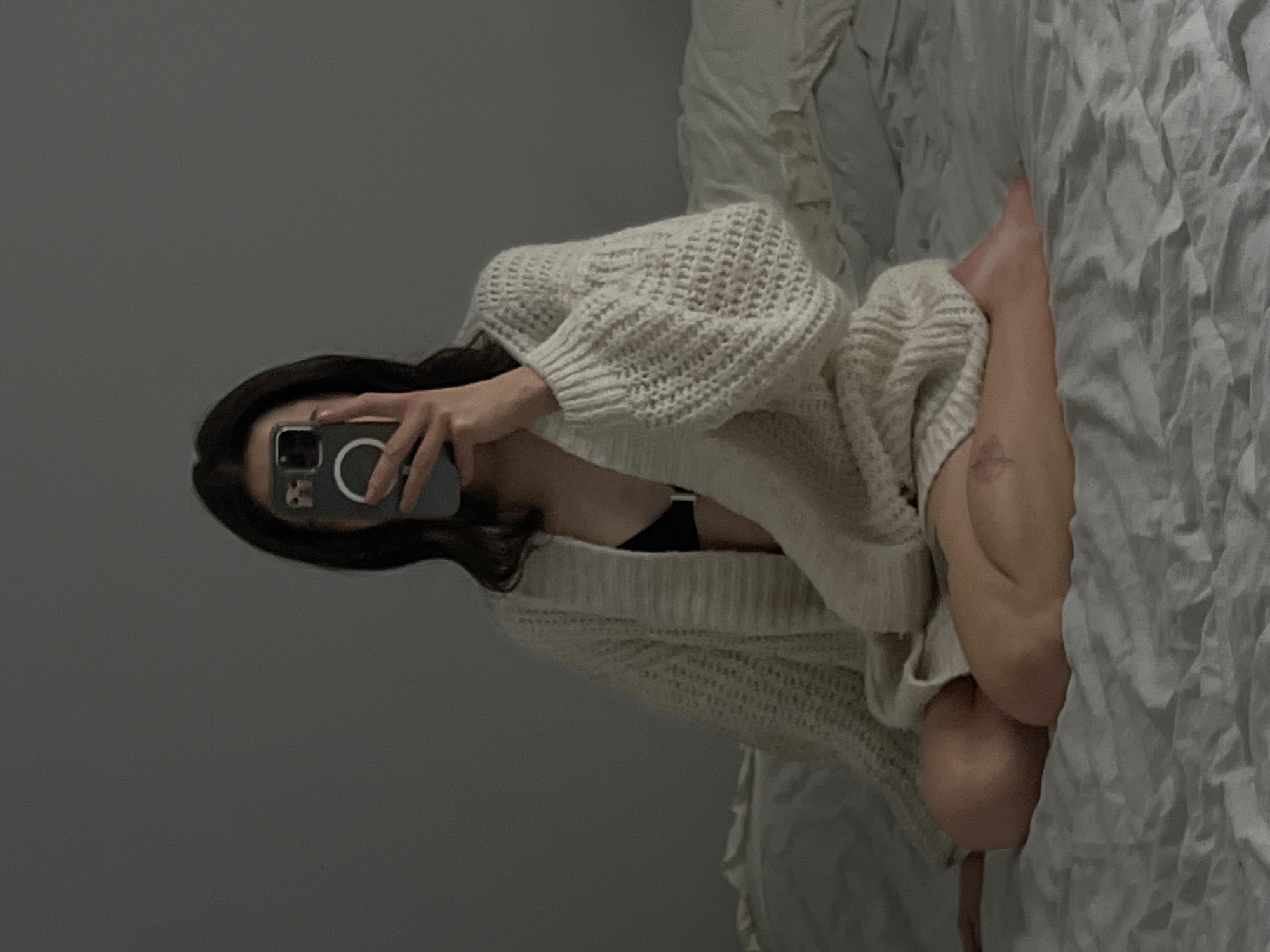 A person is sitting cross-legged on a bed, taking a mirror selfie with their phone. They are wearing a black bra and an oversized cream-colored knitted cardigan. The background is a plain gray wall. The lighting is dim, suggesting an indoor setting, possibly a bedroom. The image does not contain any information about the location in Ternopil, Ukraine.