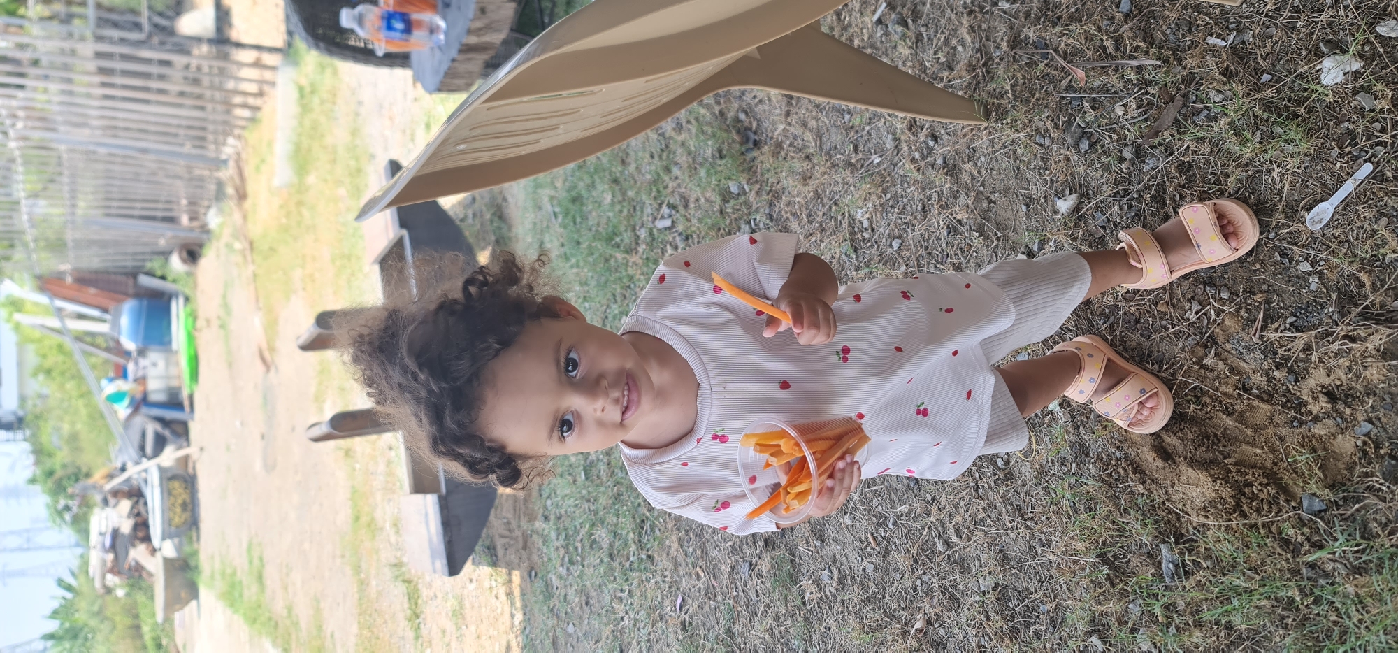 A child is standing on a dirt-covered ground. The child is wearing a white pajama with red cherry prints, and pink sandals. The child is holding a plastic cup filled with orange objects that may be carrots. A beige plastic chair and part of a wooden bench are also in view. In the background, there are trees, a fence, a blue bucket and other objects. It is not possible to accurately determine if this image was taken in Đà Nẵng, Vietnam.