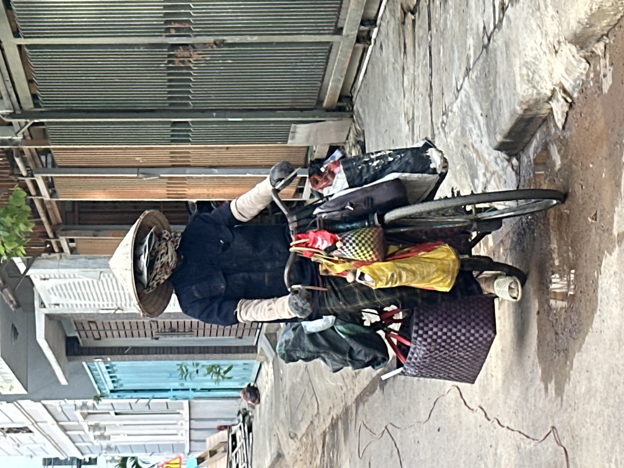 In Đà Nẵng, Vietnam, a person wearing a conical hat, dark shirt, plaid pants, and light-colored slippers is riding a bicycle laden with several bags. The person's face is obscured by a patterned scarf and part of the hat. The bicycle has bags of various colors and patterns attached to its handlebars and frame. The scene takes place on a paved street with some puddles, suggestive of recent rain. In the background, there is a building with white shutters and a blue gate, and further back, a tall metal gate. The overall impression is that of a person engaged in daily activities or work in a residential or commercial area of the city.