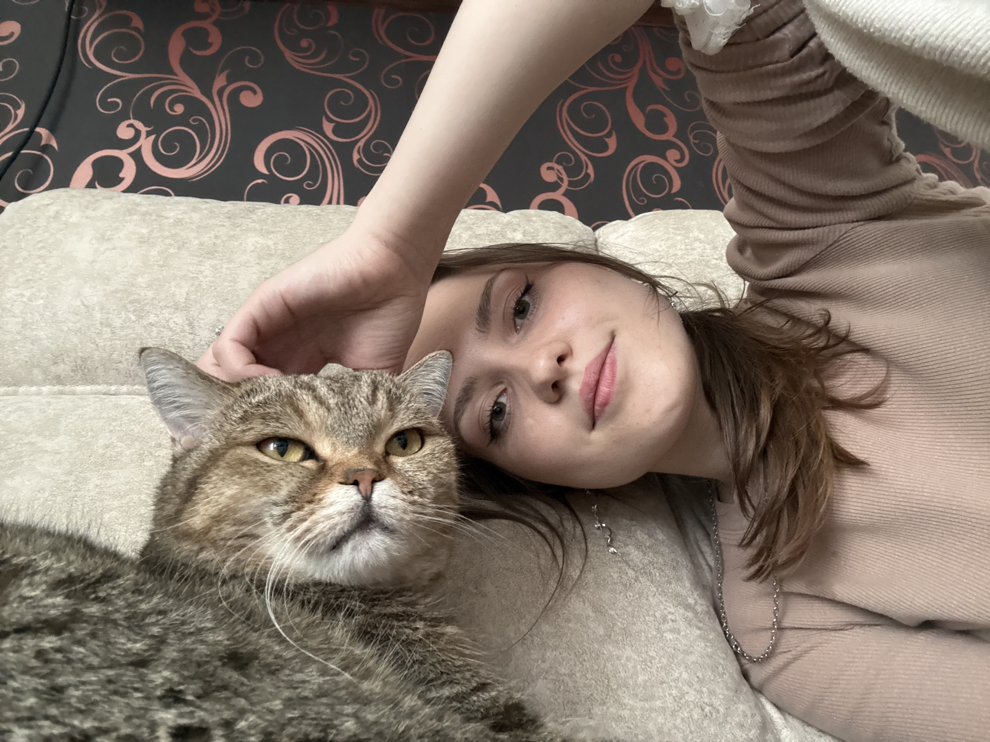 An indoor, eye-level shot shows a young woman and her cat resting together on a light-colored sofa. The woman, appearing to be in her late teens or early twenties with light brown hair, is positioned on the right, facing the camera with her head tilted and a soft smile. She wears a light beige or tan ribbed long-sleeved top and a thin silver-colored chain necklace. Her hand is gently placed on the head of the cat, suggesting affection.
The cat, a tabby with brown, black, and possibly some lighter fur, occupies the lower-left side of the frame, looking forward with its visible yellow/amber eye slightly narrowed, giving it a calm or contemplative expression. It has prominent whiskers.
In the background, a dark wallpaper with a repeating pattern of stylized pinkish-red floral or swirl motifs is visible. The scene conveys a sense of warmth and companionship between the woman and her pet. The lighting appears to be ambient indoor light. No text is visible in the image. The setting is consistent with a home environment in Saint Petersburg, Russia.