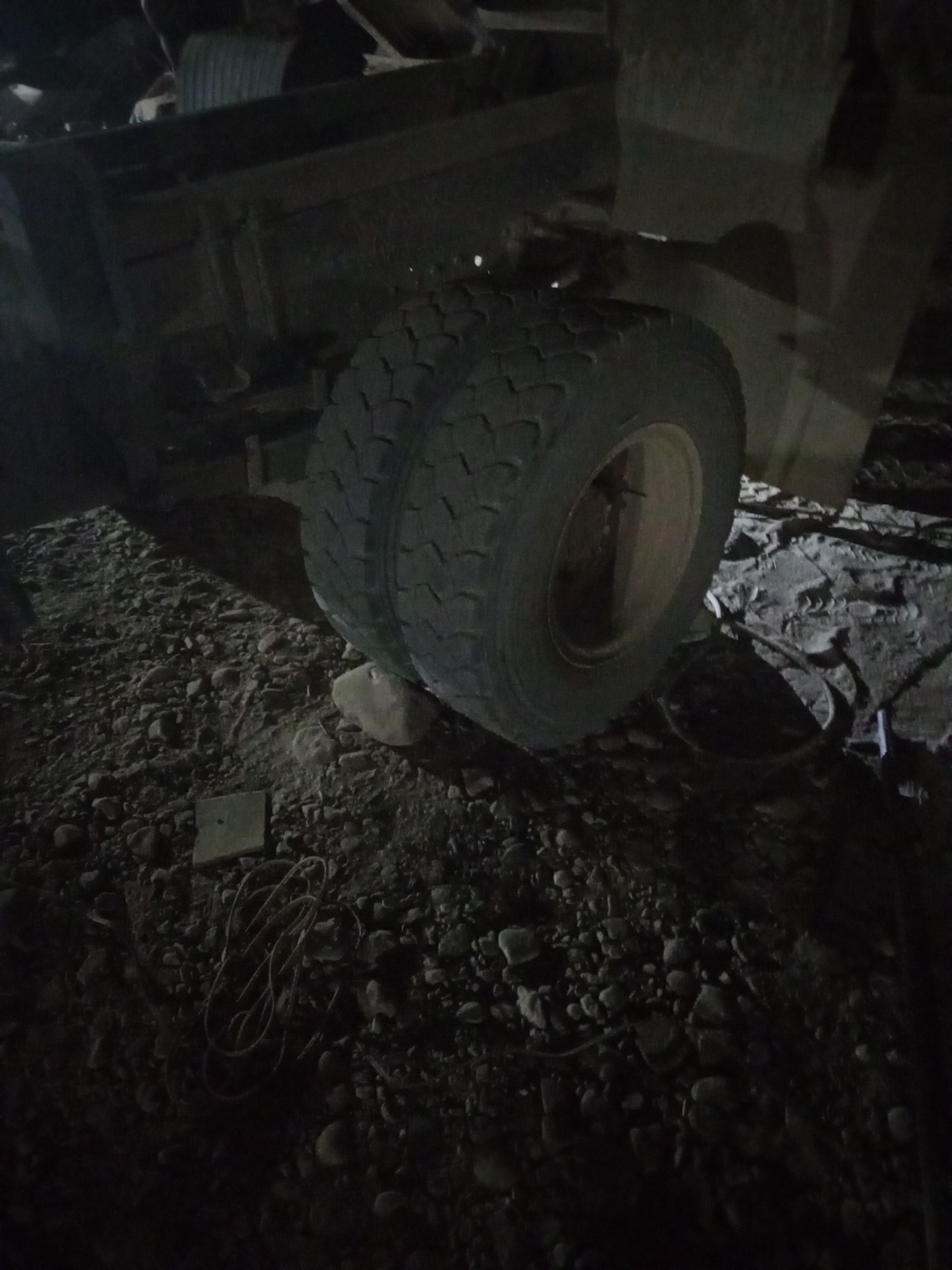 The image depicts a close-up, low-angle view of the rear dual tires of a truck at ground level in Keling Panggau, Indonesia. The tires are large and have a heavy-duty tread pattern. They are positioned on a surface covered with gravel and small rocks, with some larger stones also visible. A dark, shadow-drenched truck bed or chassis is visible above the tires. The lighting is dim, suggesting it is nighttime or a poorly lit area, with shadows cast across the ground. Some electrical wire or cable is coiled on the ground in the lower portion of the frame, near a flat, square object. The overall scene appears to be an outdoor, potentially industrial or construction site, given the presence of heavy machinery and the rough terrain. There are no people visible in the image.