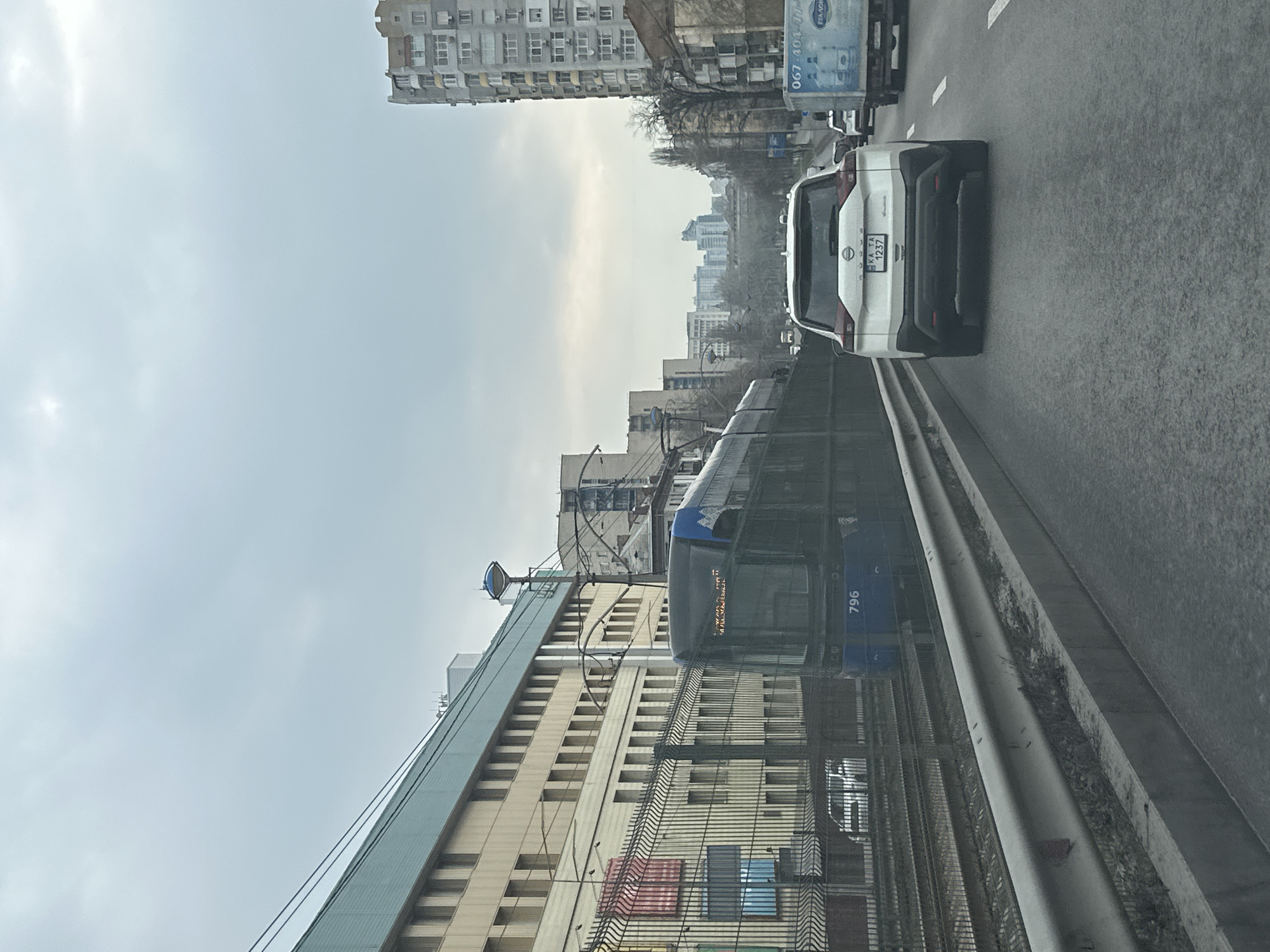 The image captures an urban scene in Kyiv, Ukraine. A white Nissan Rogue with license plate KKA TA 1237 is traveling on a wet road, likely a highway or multi-lane street, with other vehicles present. Adjacent to the road, separated by a guardrail and fencing, is a tram line. A blue tram, marked with the number 796, is traveling on the tram line. Beyond the tram line, there is a large building with multiple windows. In the background, other buildings are visible, including a tall apartment building and other structures that make up the Kyiv skyline. The sky is overcast, suggesting a cloudy or rainy day. The environment appears to be a mix of residential and transportation infrastructure.