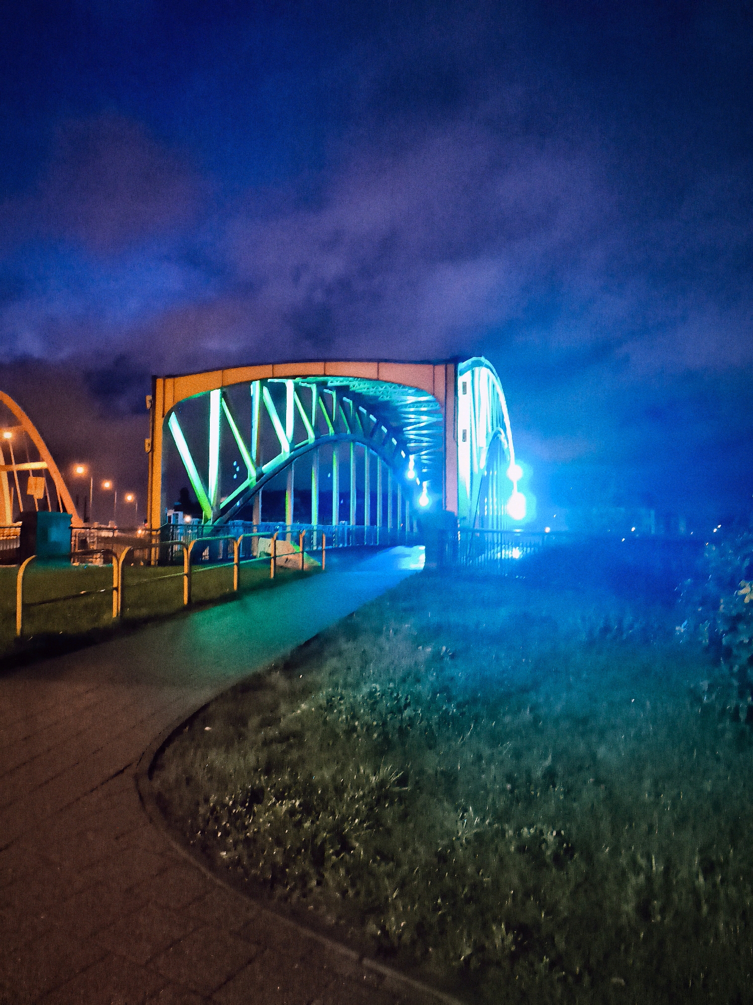 The image shows the Most Grunwaldzki bridge in Gdańsk, Poland, at night. The bridge is illuminated with green and blue lights, casting a colorful glow on the surrounding area. The sky is dark blue with some clouds. A paved walkway curves around a grassy area in the foreground. The bridge itself is a steel arch bridge with a distinctive lattice structure. The lighting creates a dramatic and somewhat surreal atmosphere. There are no people clearly visible in the image, though a dark silhouette of a person can be seen on the bridge. The scene appears to be peaceful and quiet, with no signs of activity other than the illumination of the bridge.