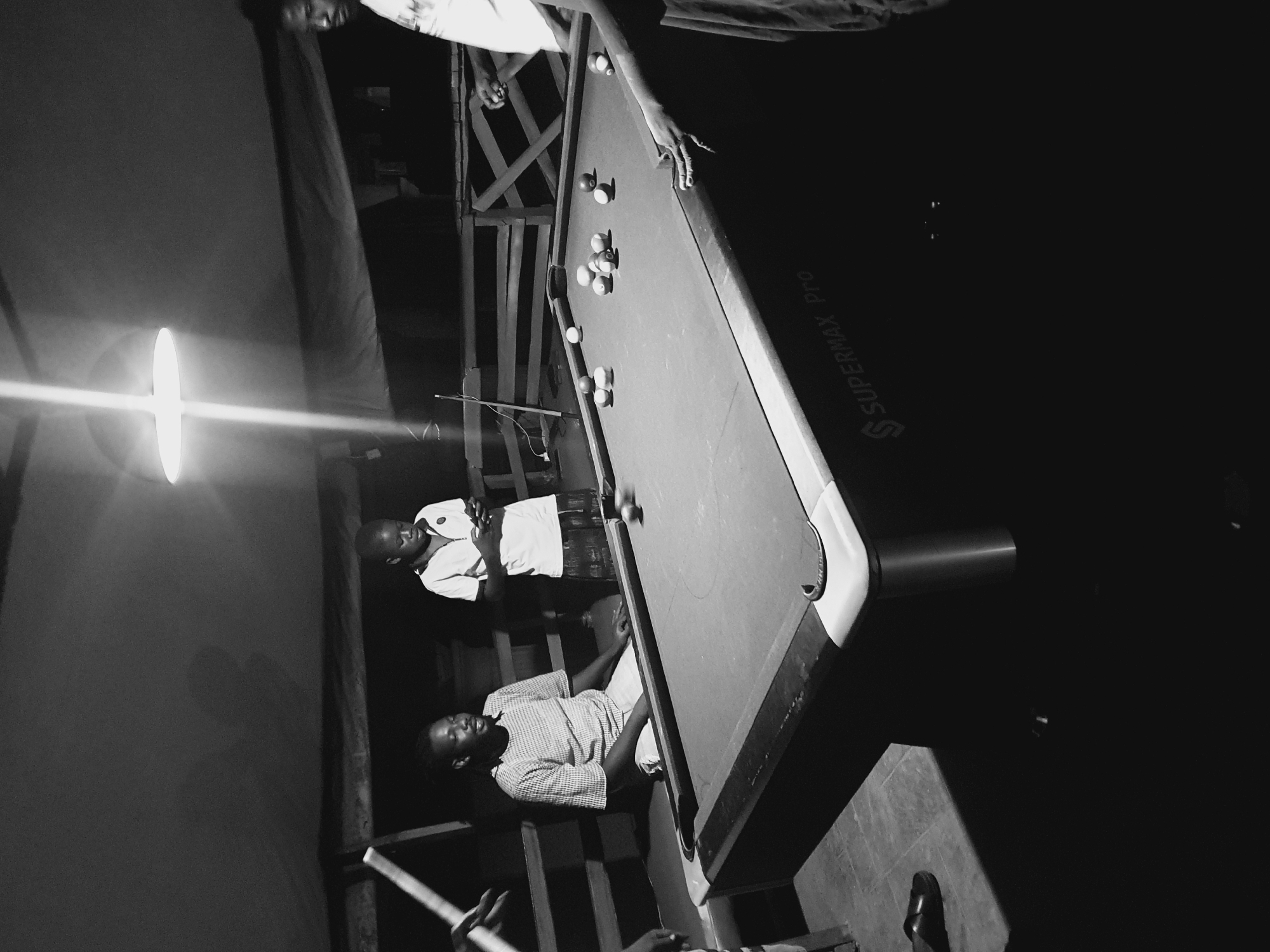 The black and white photograph captures a group of individuals engaged in a game of pool in a dimly lit outdoor setting. The scene appears to be an open-air bar or recreational area, possibly located in Lokoja, Nigeria, given the context provided. A pool table, marked with the "SUPERMAX Pro" branding on its side, occupies the center of the frame, laden with several pool balls. Four people are visible around the table.
On the left, a man in a checkered shirt is seated, looking towards the table. To his right, a younger individual in a white polo shirt is standing, engrossed in their phone. Further to the right, another person's hands are visible near the edge of the pool table, suggesting they are either playing or observing the game. In the background, wooden fencing and what appears to be a tarp or canopy create the boundary of the space. A bright overhead light fixture casts a strong illumination downwards, creating stark contrasts and shadows. The floor is tiled. The overall impression is one of casual recreation and social gathering in a humid, tropical environment.