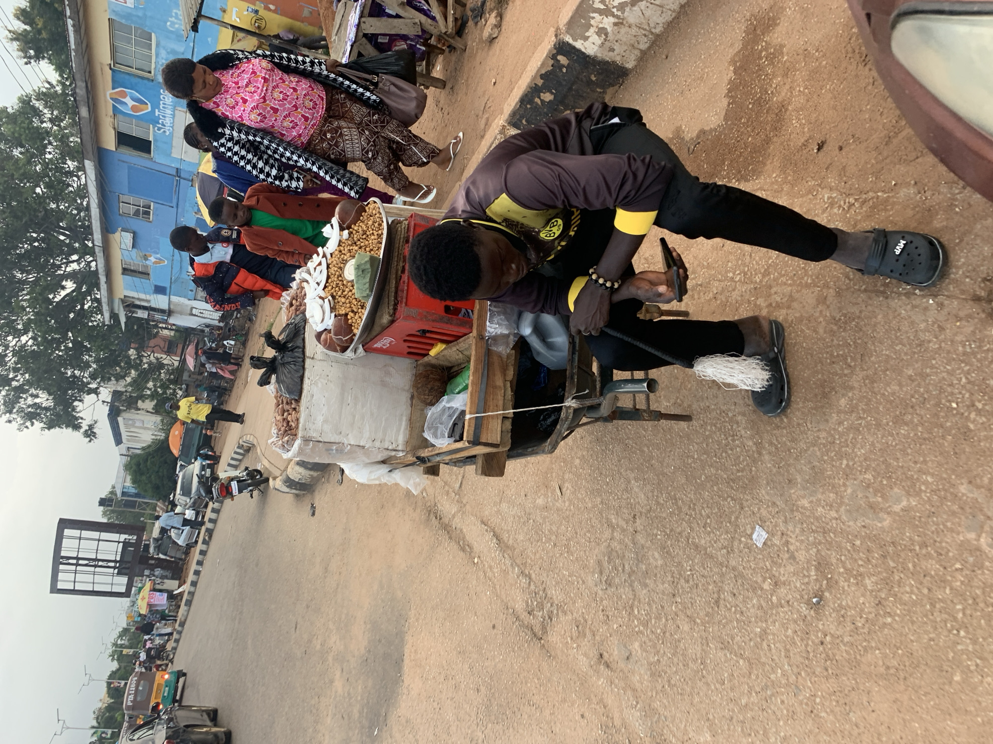 In Jos, Nigeria, on a dusty, paved roadside, a young man wearing a dark long-sleeved shirt with yellow accents and black pants sits on a cart. He is holding a black smartphone in his right hand, looking down at it. A mop with white strings hangs from the cart near his feet. The cart itself is laden with various goods, including bags of nuts, coconuts, and other food items displayed in a metal tray. Behind him, a woman in a pink and white patterned top and patterned trousers, wearing a black and white patterned coat, walks away, holding a dark bag. Further in the background, other people are visible, some standing and some walking, near a blue and white building with the "StarTimes" logo. Several vehicles, including a tricar, a motorcycle, and cars, are present on the road. The environment suggests a busy market or roadside vendor area.
