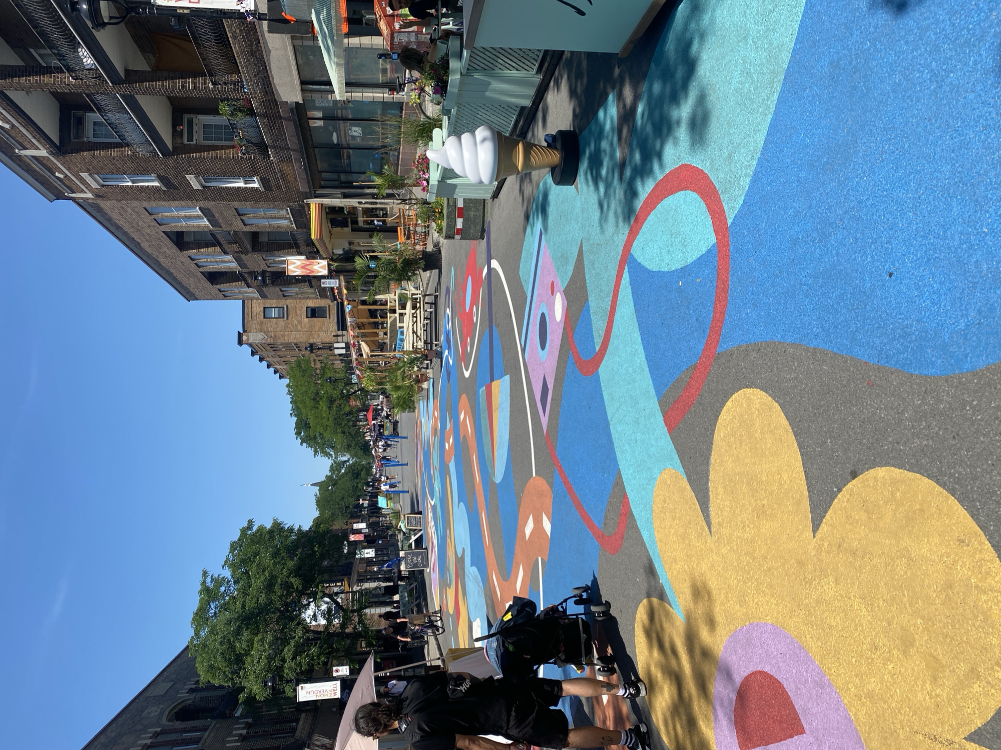 The image shows a vibrant street art installation in Montreal, Canada. The asphalt has been painted with a colorful, abstract design featuring large, flowing shapes in blue, yellow, purple, red, and orange. This artwork covers the entire width of the street, suggesting it is a pedestrianized area or a street closed for an event. In the foreground, a person wearing a black t-shirt and shorts, with a backpack, stands next to a stroller. They are walking on the painted street.
On the sides of the street, there are brick buildings with multiple stories, balconies, and businesses at street level, including cafes with outdoor seating and awnings. Trees line the sidewalks, providing shade. Further down the street, more people are visible walking and cycling, enjoying the sunny weather and the festive atmosphere. In the mid-ground, there is a large, realistic sculpture of an ice cream cone. The sky is clear and blue, indicating a bright, summer day. The overall impression is of a lively, community-oriented urban space transformed by art.