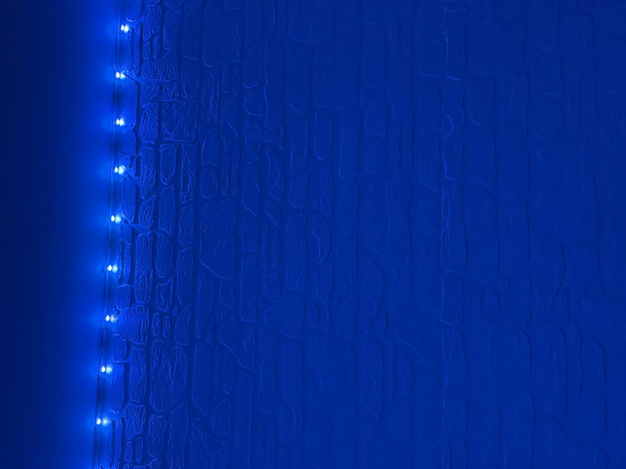 A close-up shot displays a textured blue wall, with a string of illuminated blue lights positioned along the left edge. The wall's texture resembles irregular stone or brickwork.