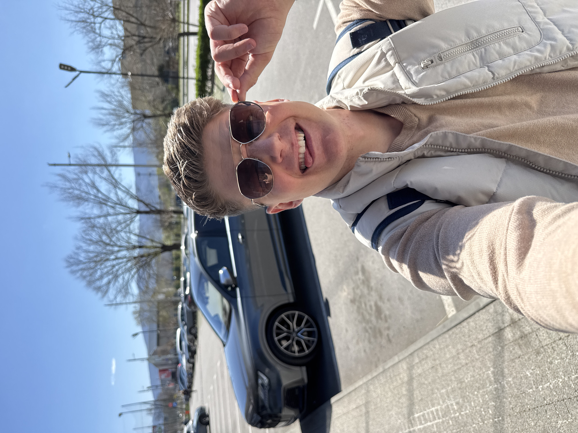 A man is taking a selfie in a parking lot. The man is light-skinned with short blonde hair, wearing sunglasses, a beige sweater, and a light-colored vest with navy blue trim. He is smiling and holding the frame of his sunglasses with his left hand. Behind him is a row of parked cars, mostly dark-colored. In the background are bare trees and a blue sky. The parking lot is paved with gray asphalt marked with white parking lines. This image was not taken in Plovdiv, Bulgaria.