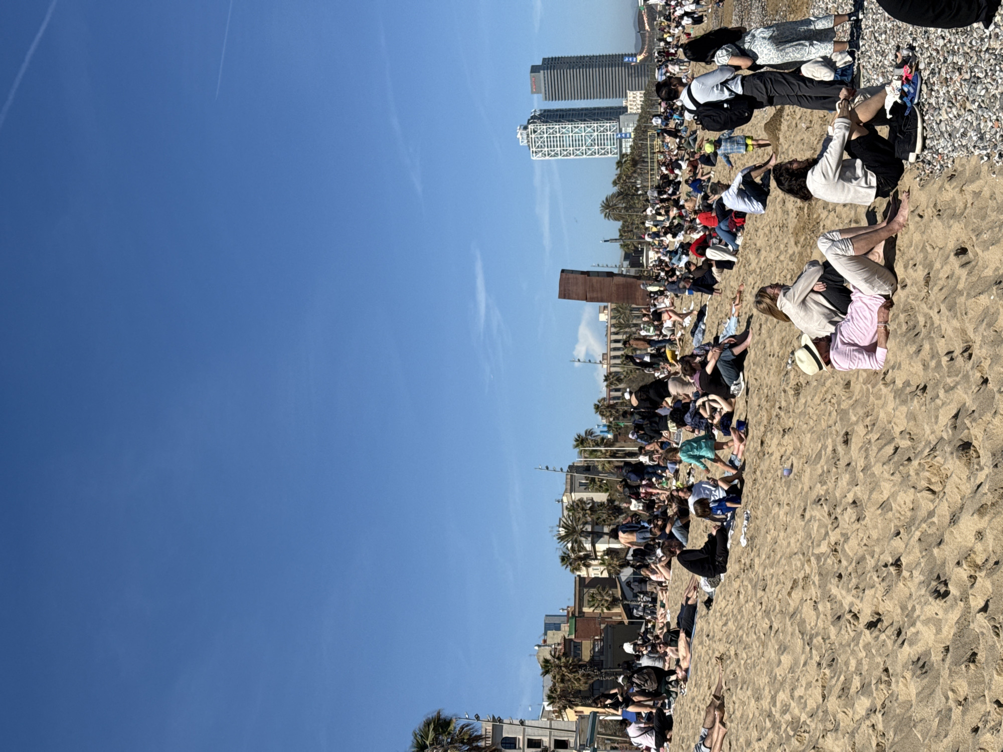 A beach scene in Barcelona, Spain, is observed under a clear, blue sky. The foreground is dominated by a sandy beach populated with numerous people. Many are lying down, while others are sitting, suggesting relaxation or sunbathing. The background reveals buildings along the coastline, including two prominent, modern towers. The architecture and palm trees indicate the location is likely a Mediterranean urban area.