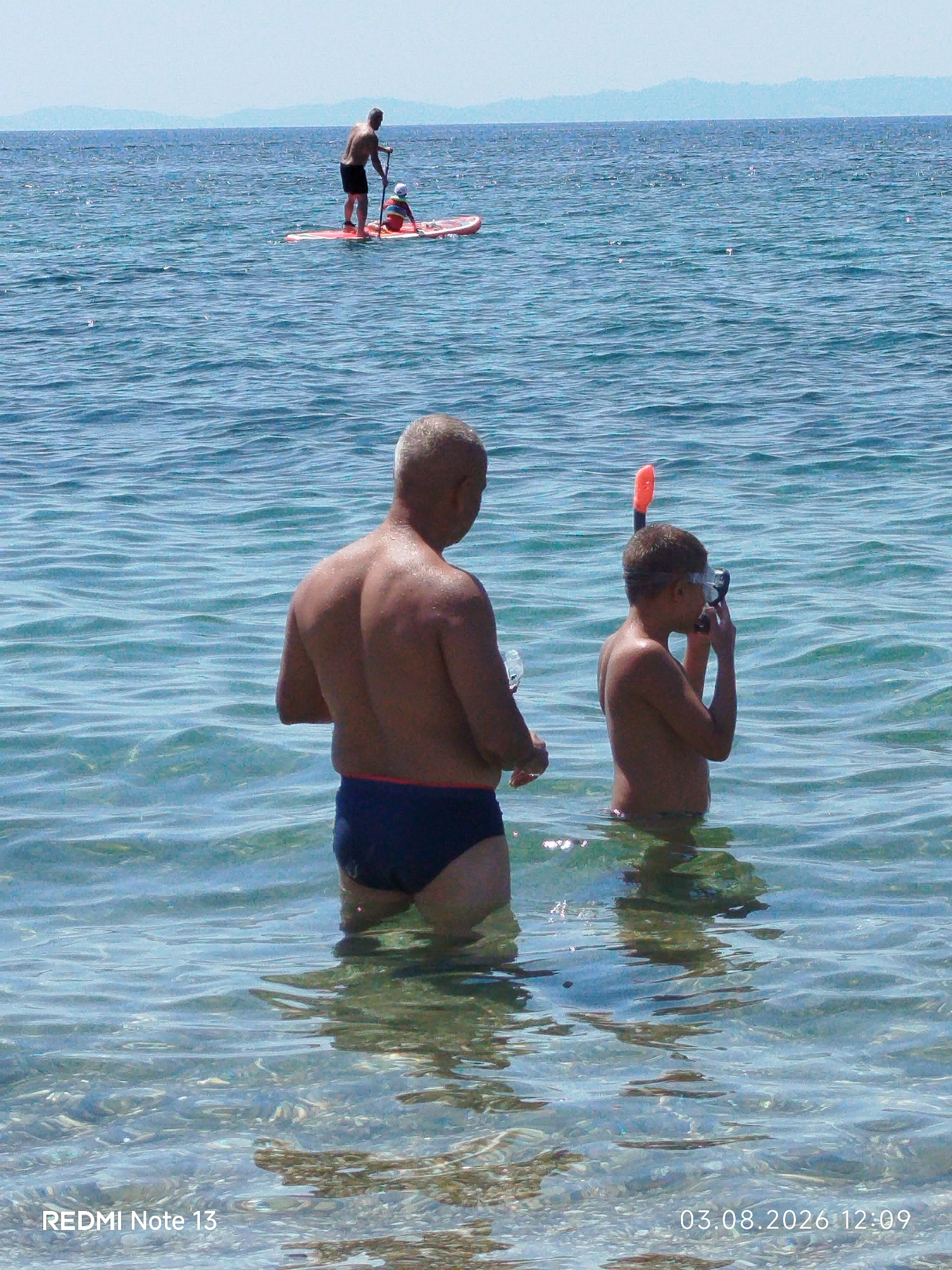 The image shows two people standing in clear, shallow ocean water. The person in the foreground on the left is an adult male, facing away from the camera, wearing dark blue swim trunks. The person on the right is a child, facing to the left, wearing a snorkel mask and an orange snorkel. In the background, a person is standing on a paddleboard with a child. The water is calm with ripples, and the seabed is visible with small rocks and pebbles. Distant hazy landmasses can be seen on the horizon under a light sky. The photograph was taken in the Sithonia Municipal Unit, Greece.