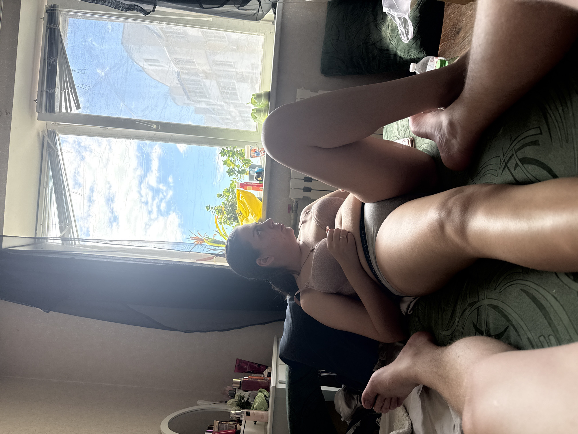 The image shows two people in what appears to be a bedroom. One person, a woman, is lying on her side, facing away from the camera, and looking towards a window. She is wearing a bra and underwear. The other person's legs are visible in the foreground and background. The scene is brightly lit, suggesting it is daytime. Sunlight streams in through a window that is partially covered by blinds and a sheer curtain. Outside the window, the sky is blue with scattered clouds, and there are buildings visible. On the windowsill, there are some items, including plants and bottles. A green patterned pillow is visible on the bed. There is some text visible on the waistband of the underwear worn by the woman, but it is difficult to read clearly. The overall mood seems relaxed and intimate.